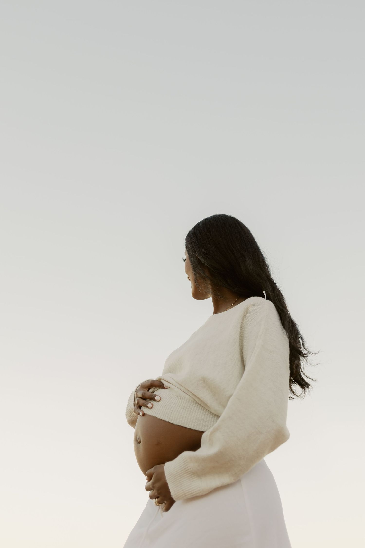 Side profile of pregnant woman in cream sweater cradling baby bump against soft grey sky background.