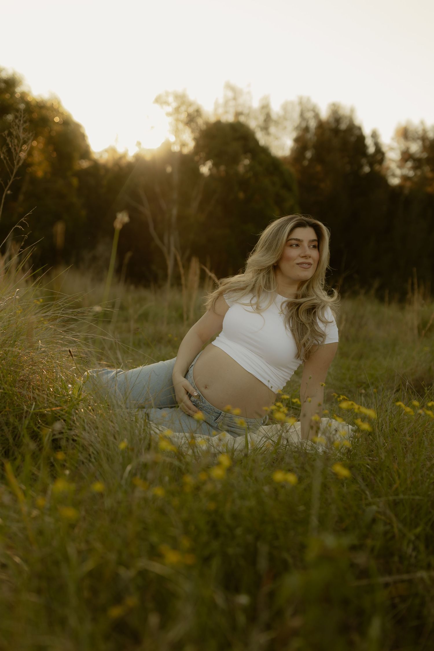 Pregnant woman in white crop top sitting in lush green field with wildflowers at sunset.