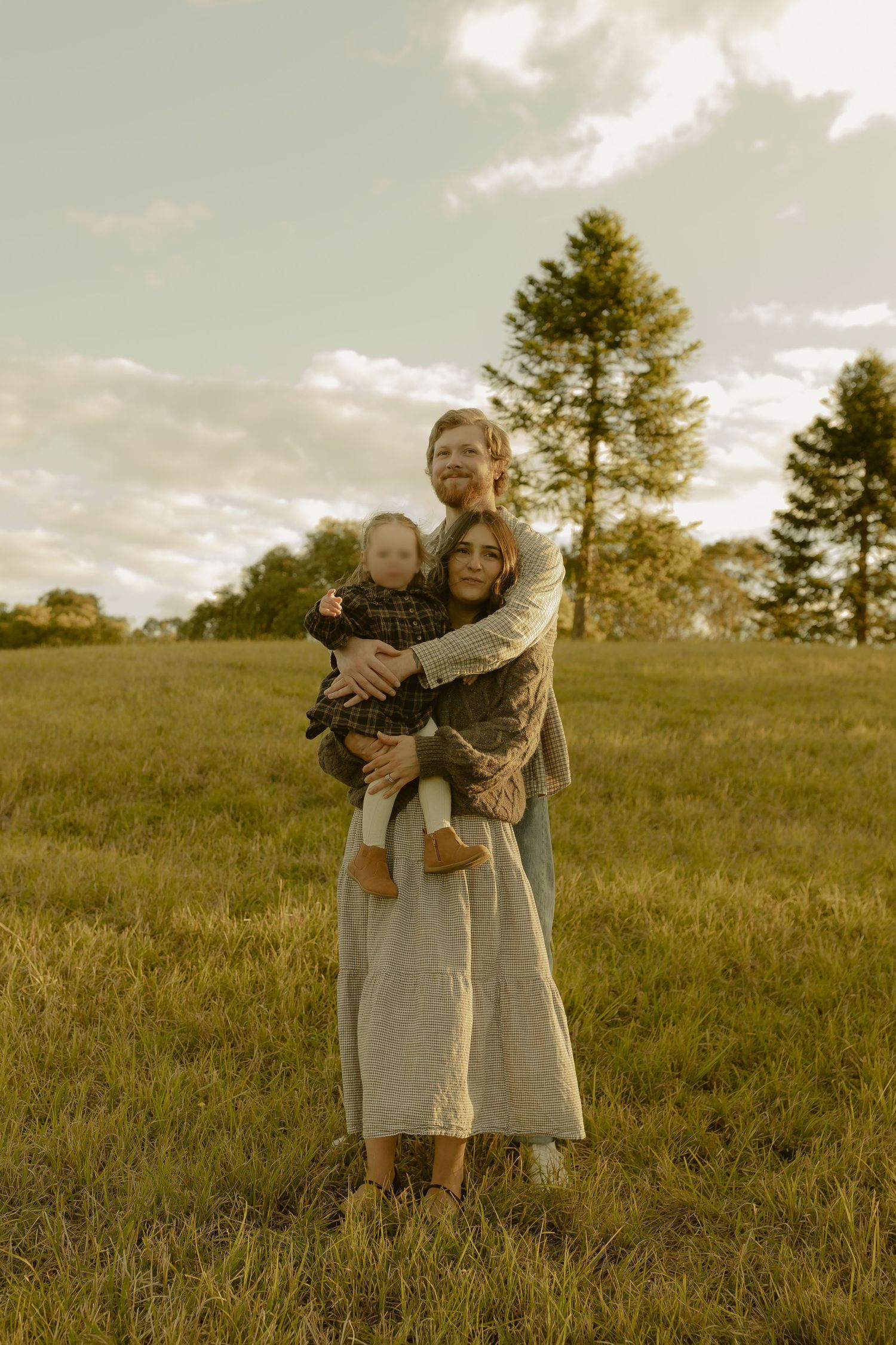 Family of three posing in a golden field at sunset, parents holding baby, trees in background.