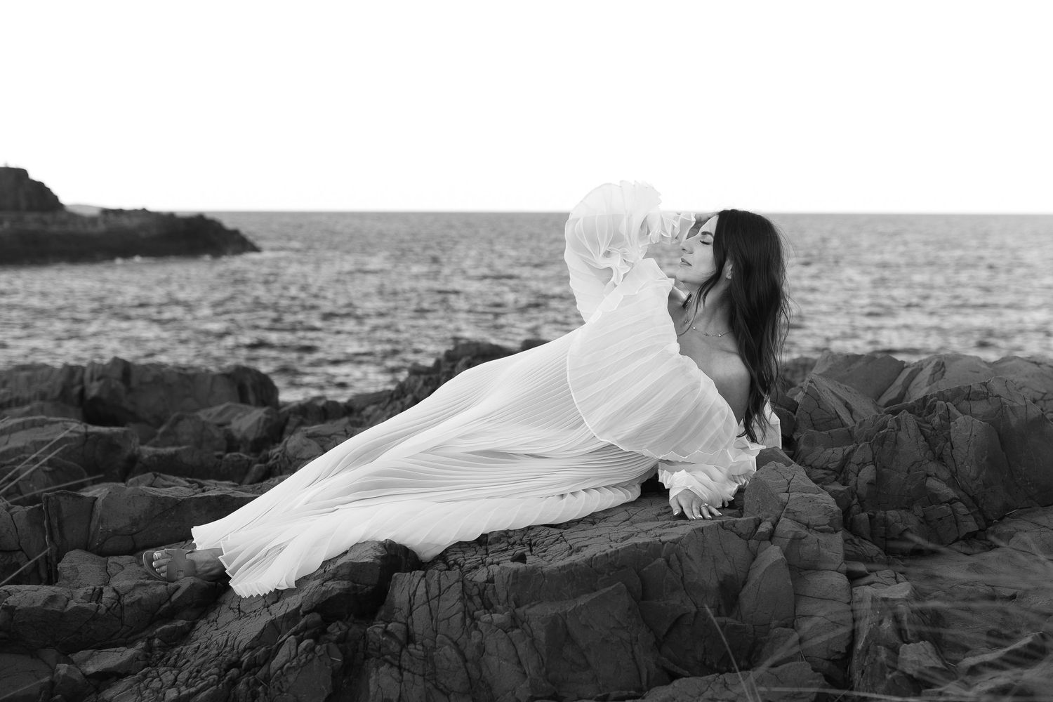 A woman in a white dress reclines on coastal rocks by the ocean in black and white photography.