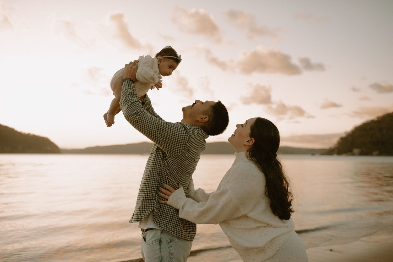 A family sharing a tender moment together on a beach at sunset with calm waters and mountains in the background.