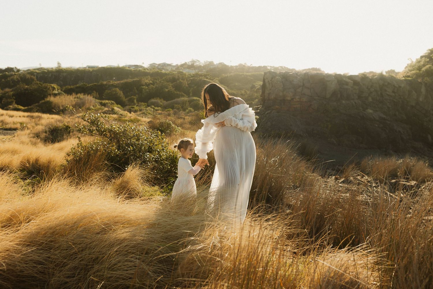 Golden light illuminates a figure in a white dress walking through tall dry grass in a rustic outdoor setting.