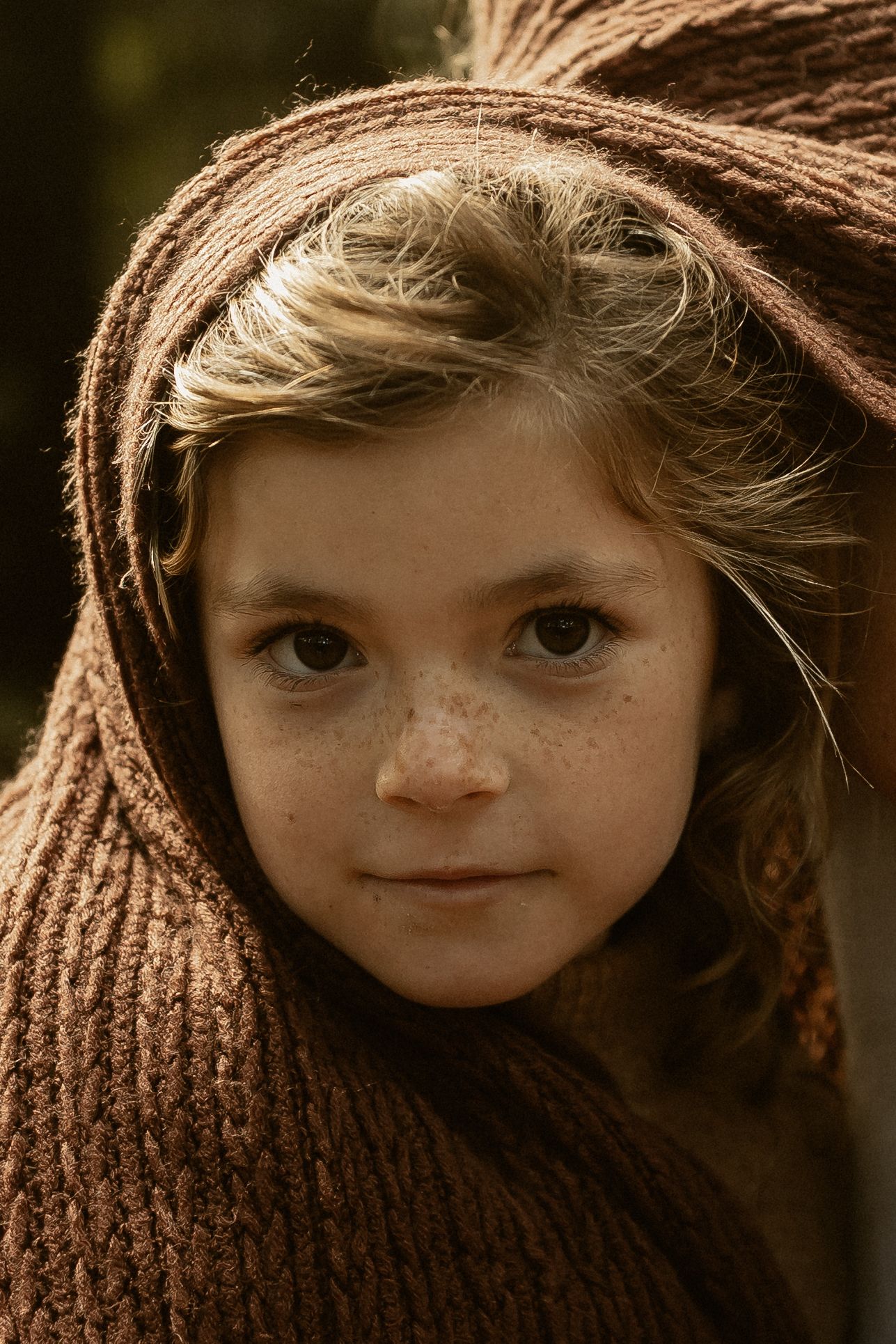 A young person peers through a cozy brown knit scarf in a moody close-up portrait with warm lighting and shallow depth of field.