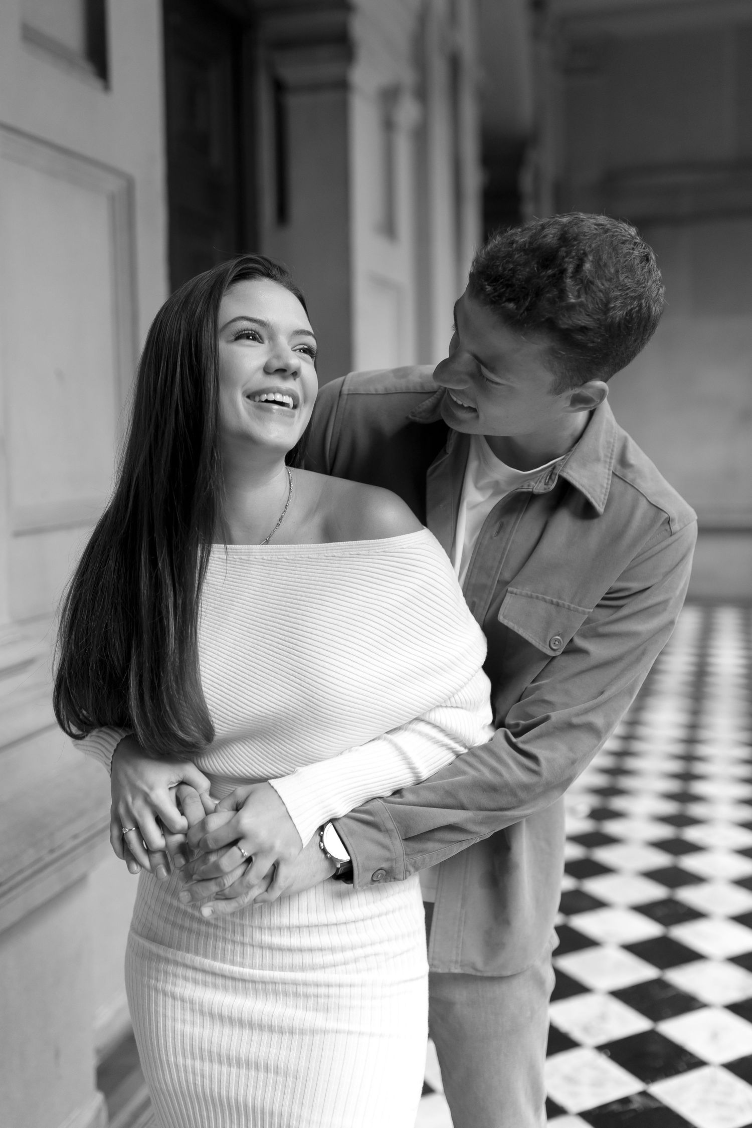 A romantic black and white photo of a couple embracing in a hallway with checkered floor tiles.