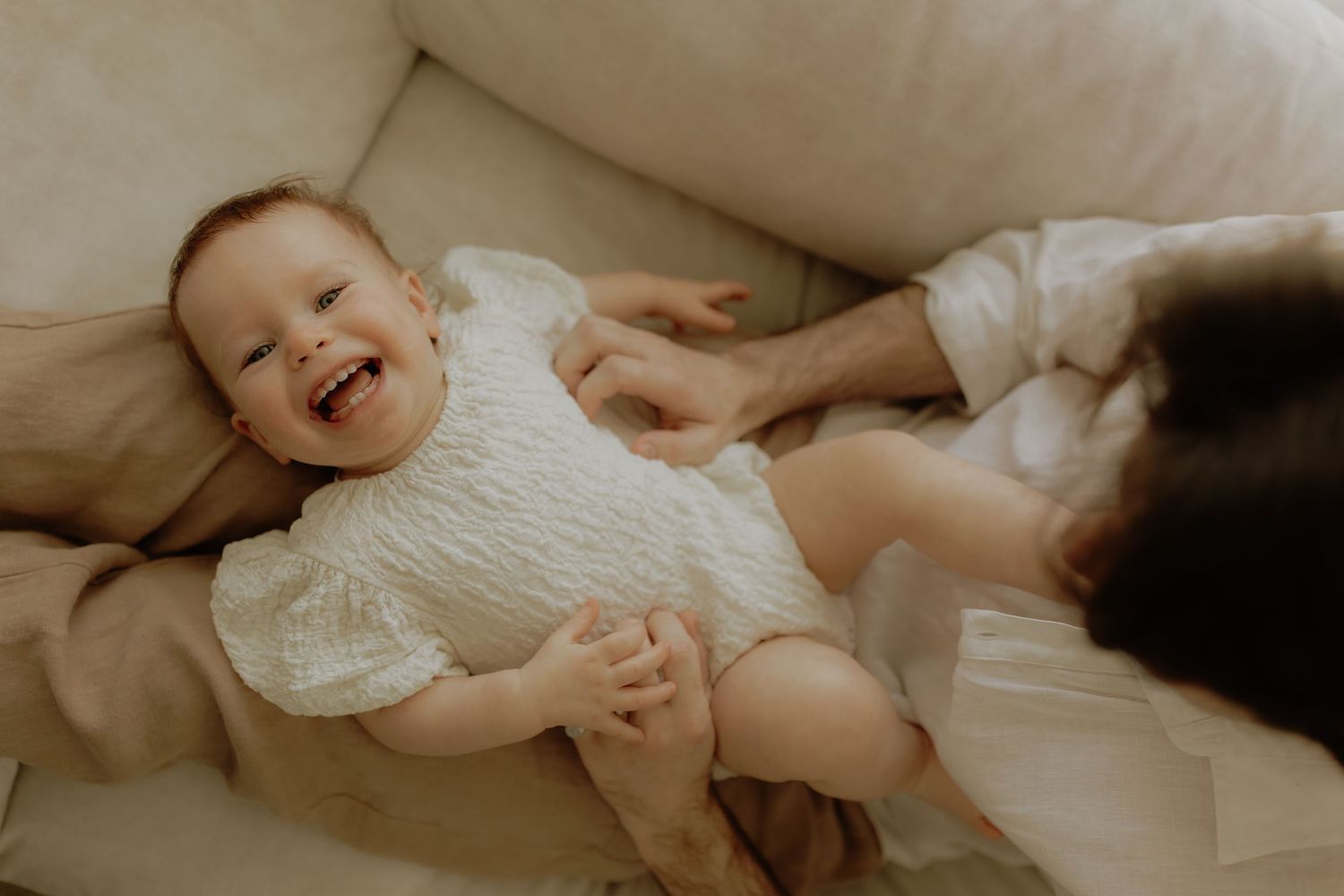 A laughing baby in a white outfit lies on a cream sofa while a parent plays with them.
