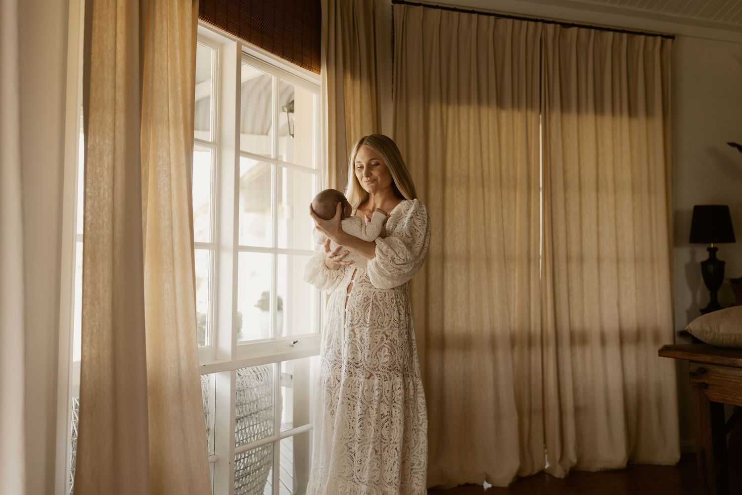 A woman in a white lace dress stands at a window with sunlight filtering through sheer curtains.