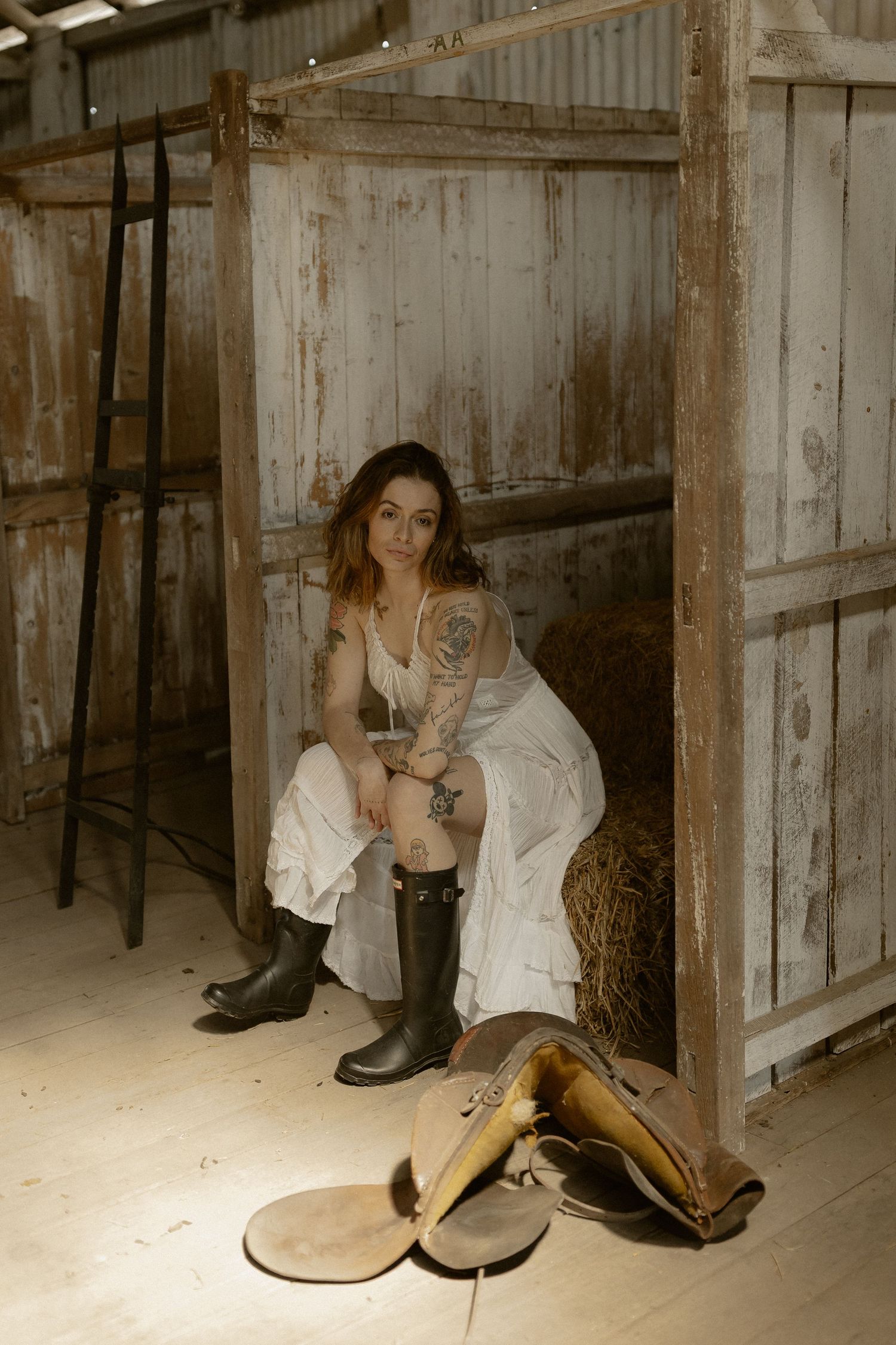White dress contrasts with rustic barn interior as boots rest nearby on hay-strewn floor.