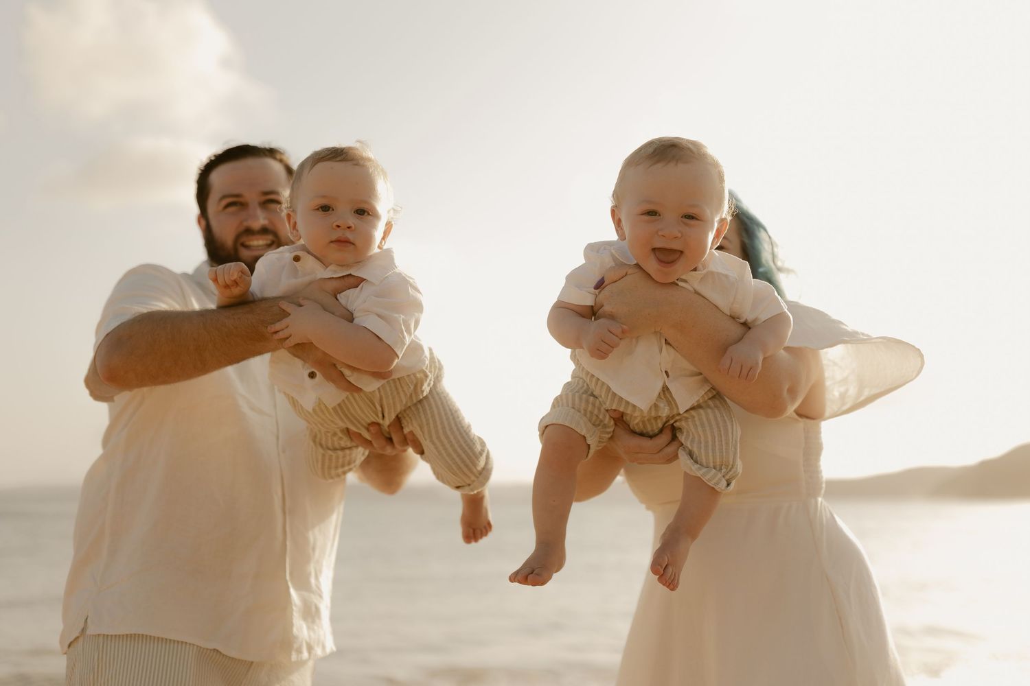 Family dressed in white holds twin babies while standing on beach at sunset.