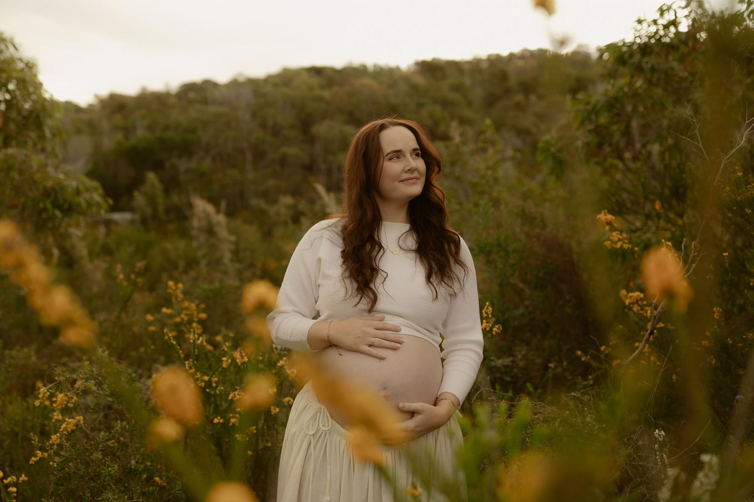 Pregnant woman in white dress cradling baby bump outdoors among wildflowers at golden hour.