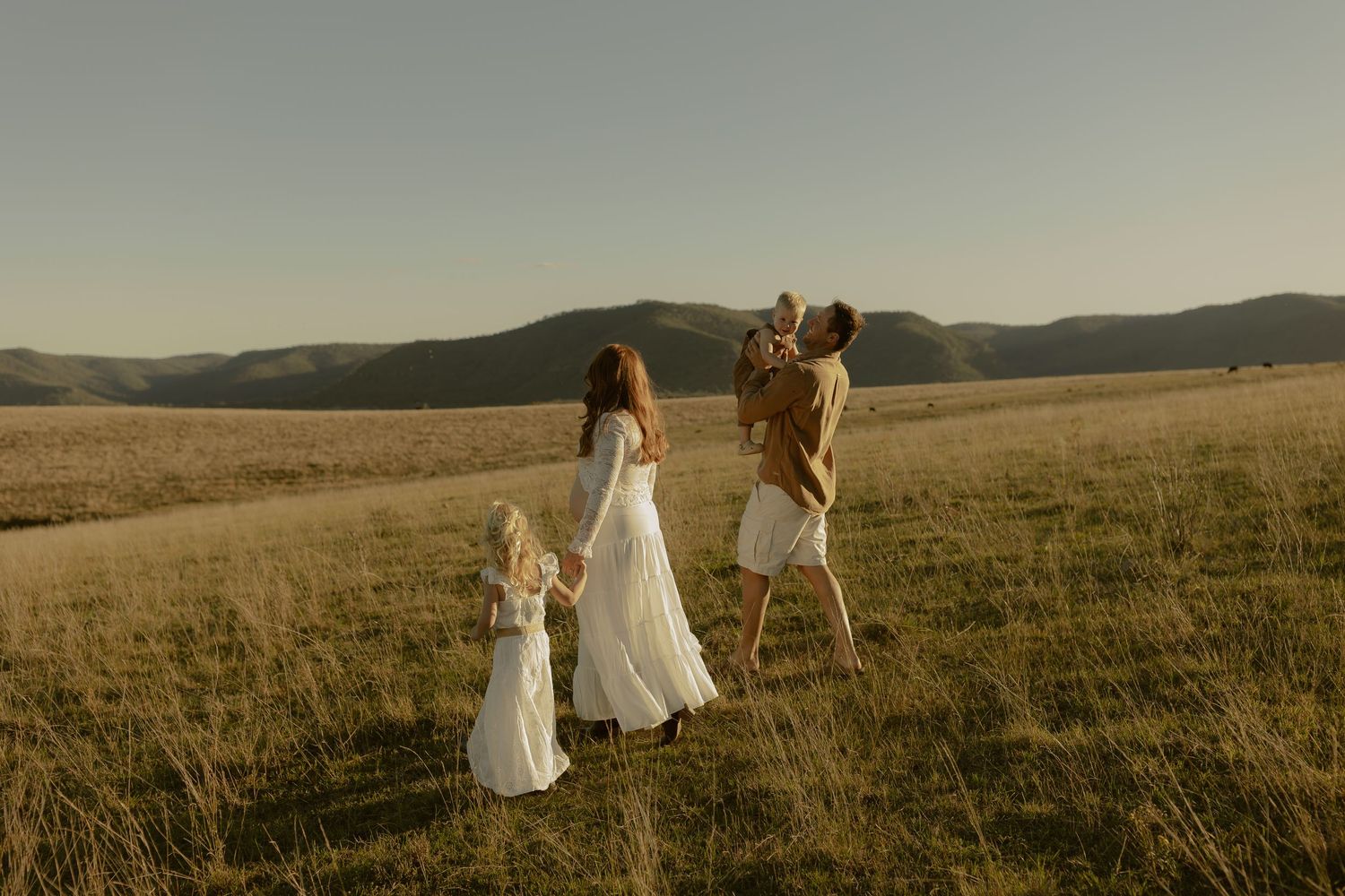 Family in white outfits walking through golden meadow with mountain backdrop in warm sunlight.