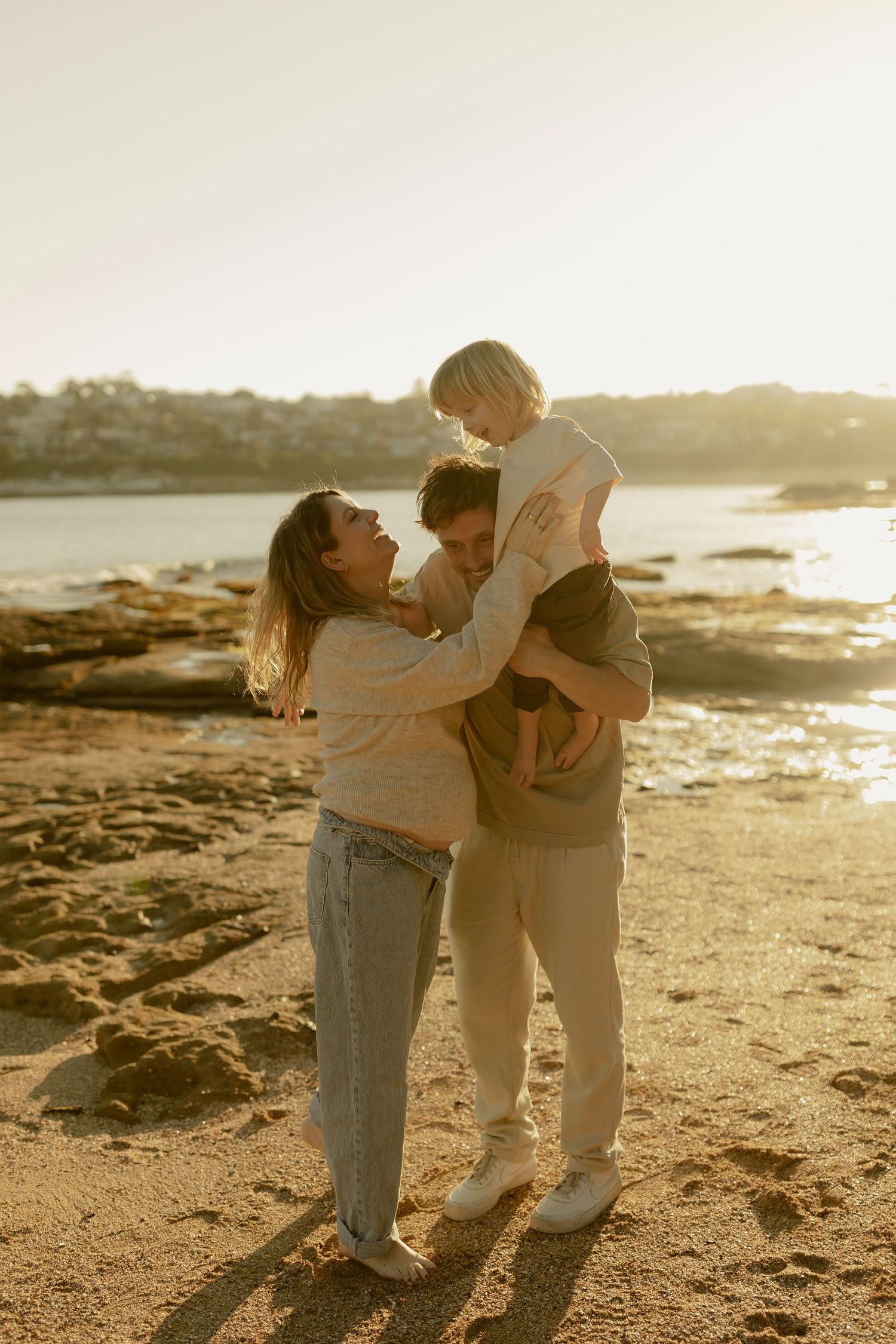 A joyful family of three shares a warm embrace on a sandy beach at golden hour sunset.