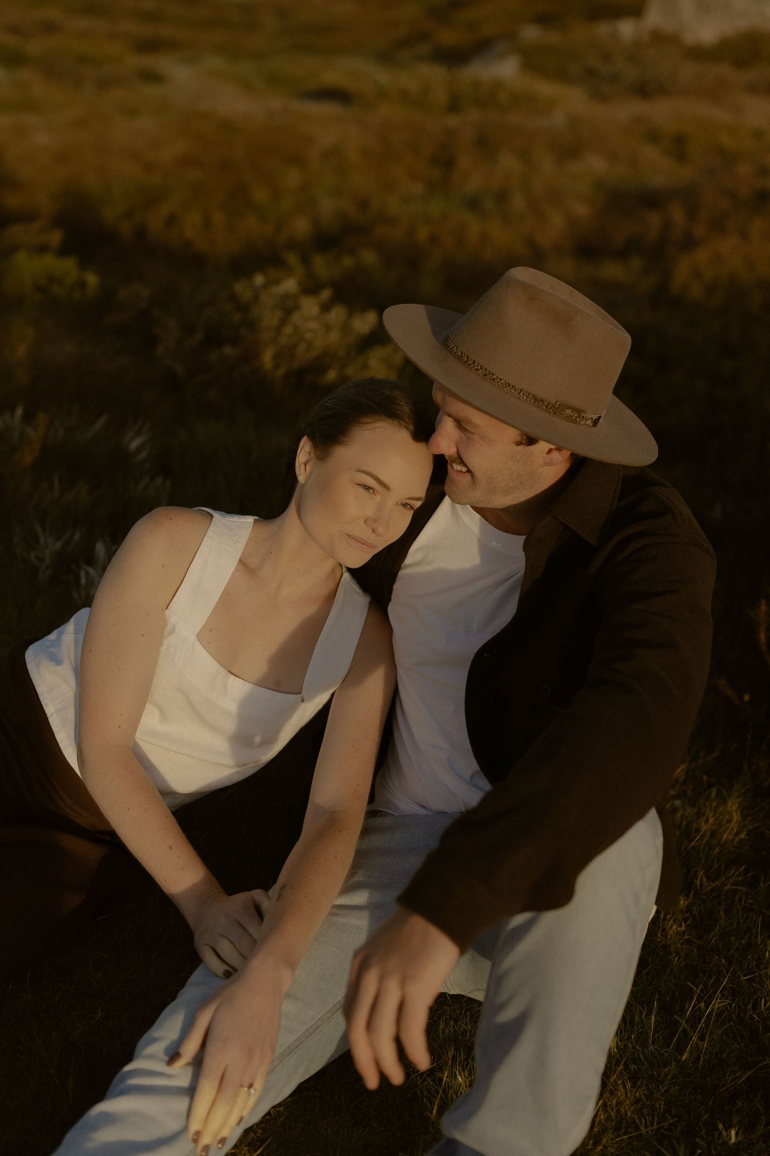 A tender moment between a couple in casual attire sitting on a grassy hillside during golden hour.