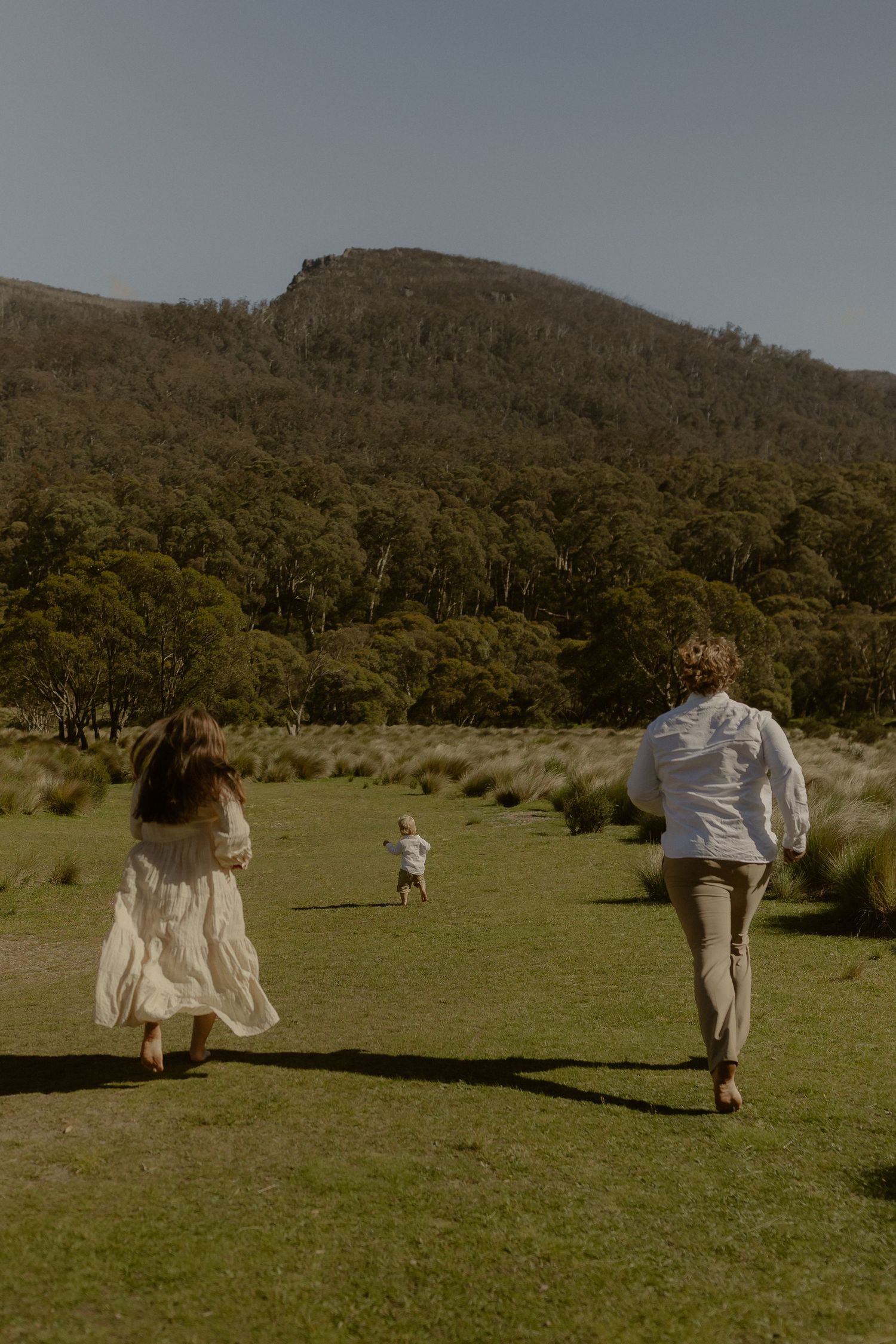 A family runs together across a green field with mountains and trees in the background.