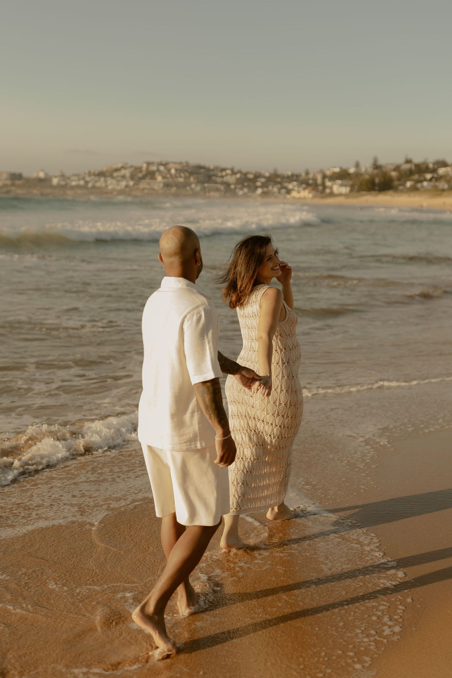 A couple walks together along a sandy beach at sunset with waves gently rolling in from the ocean.
