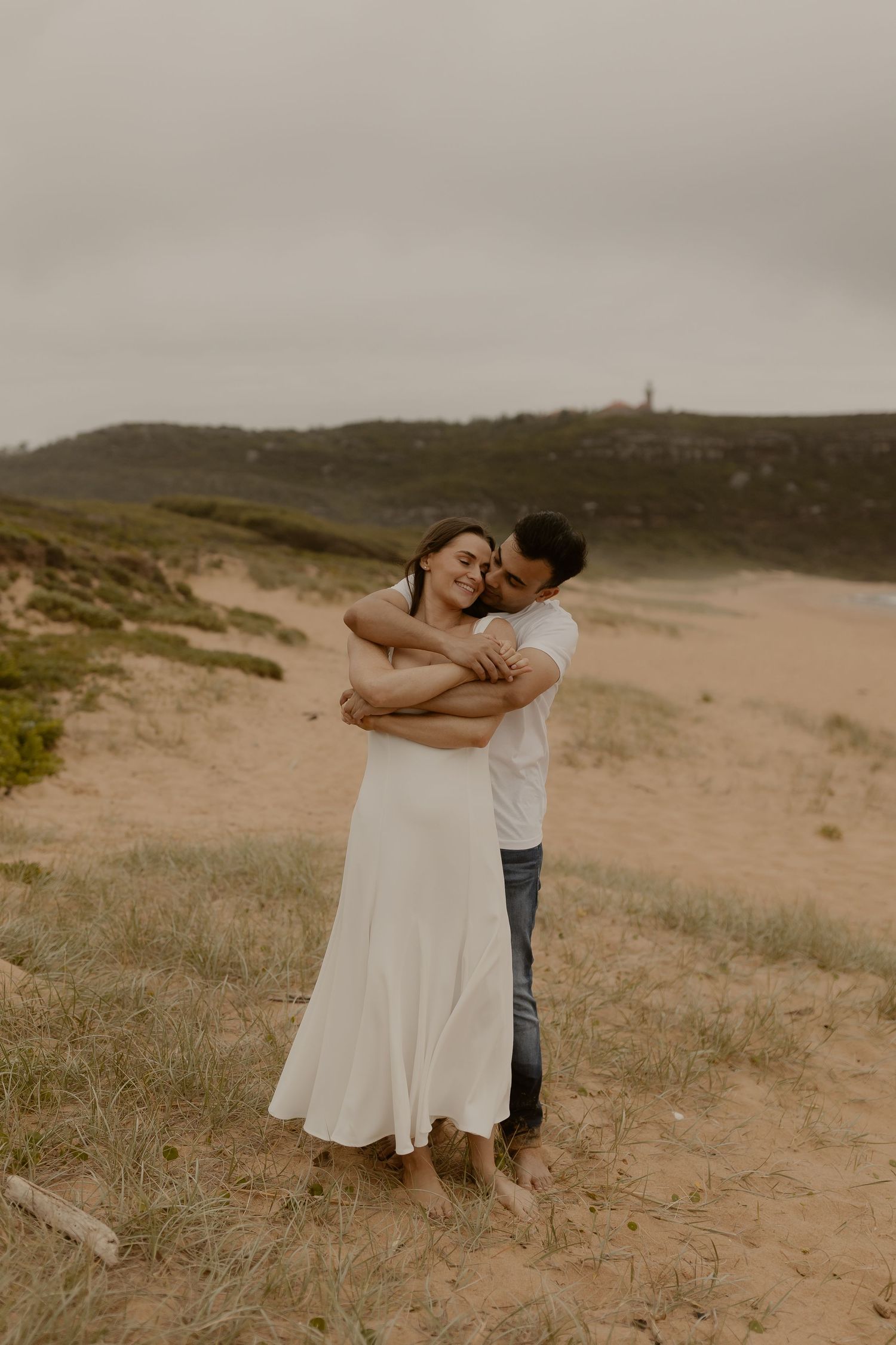 A tender embrace captured on a hilltop overlooking a scenic coastal beach at sunset.