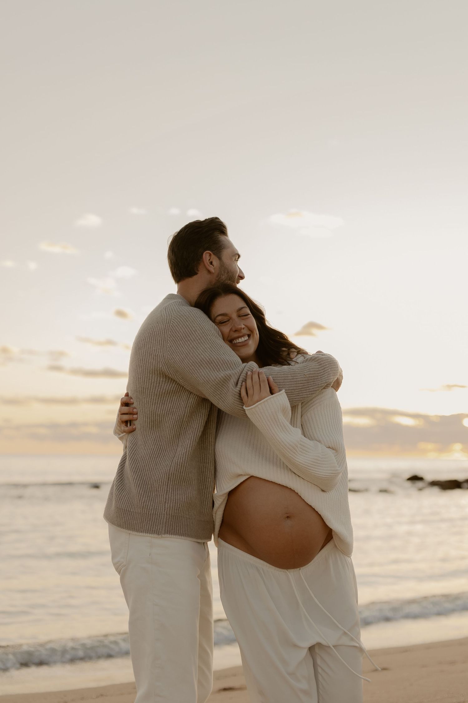 Expecting couple embraces on beach at sunset as they share a tender moment during maternity photoshoot.
