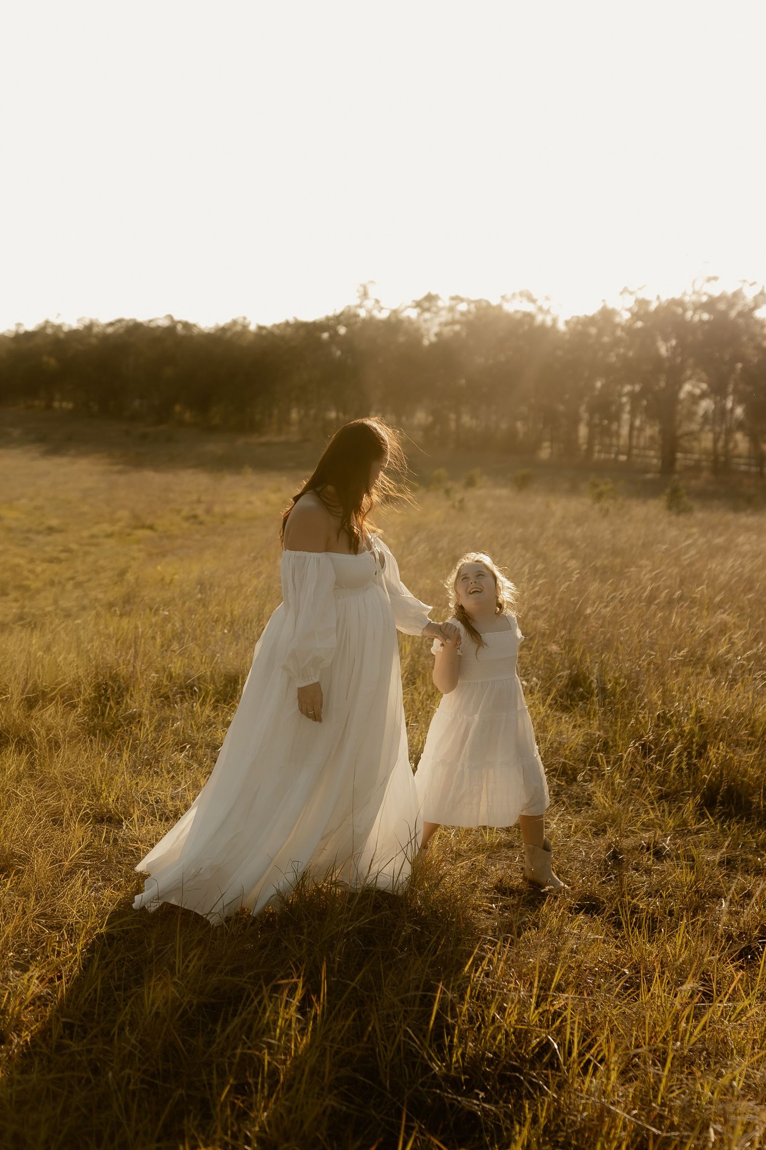 A parent and child in flowing white dresses walk through a sunlit meadow at golden hour.