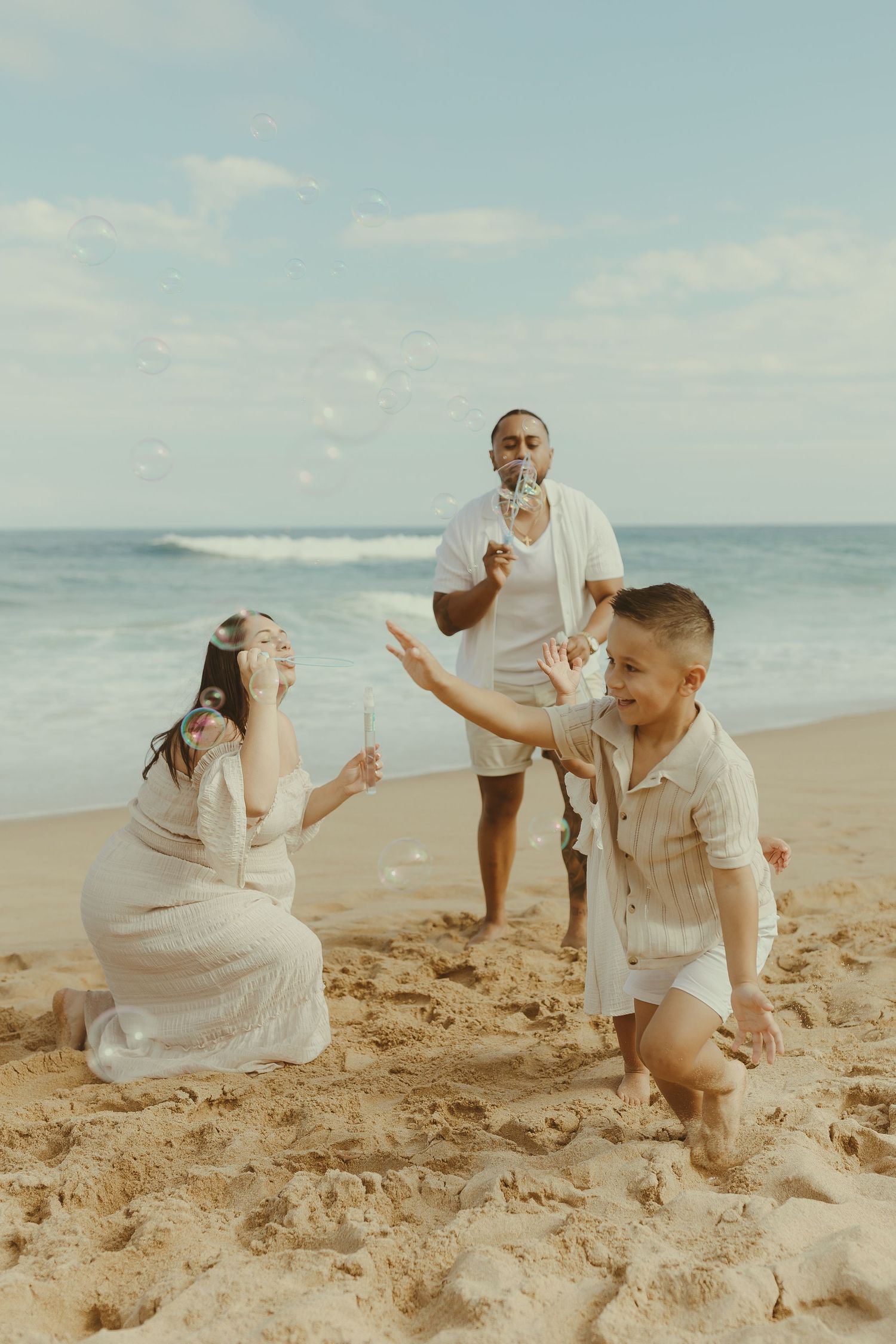 A couple in white outfits playfully poses together on a sandy beach with ocean waves in the background.