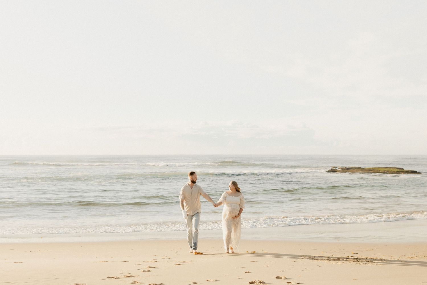 A couple in light clothing walks together along a serene beach shoreline on a misty day.