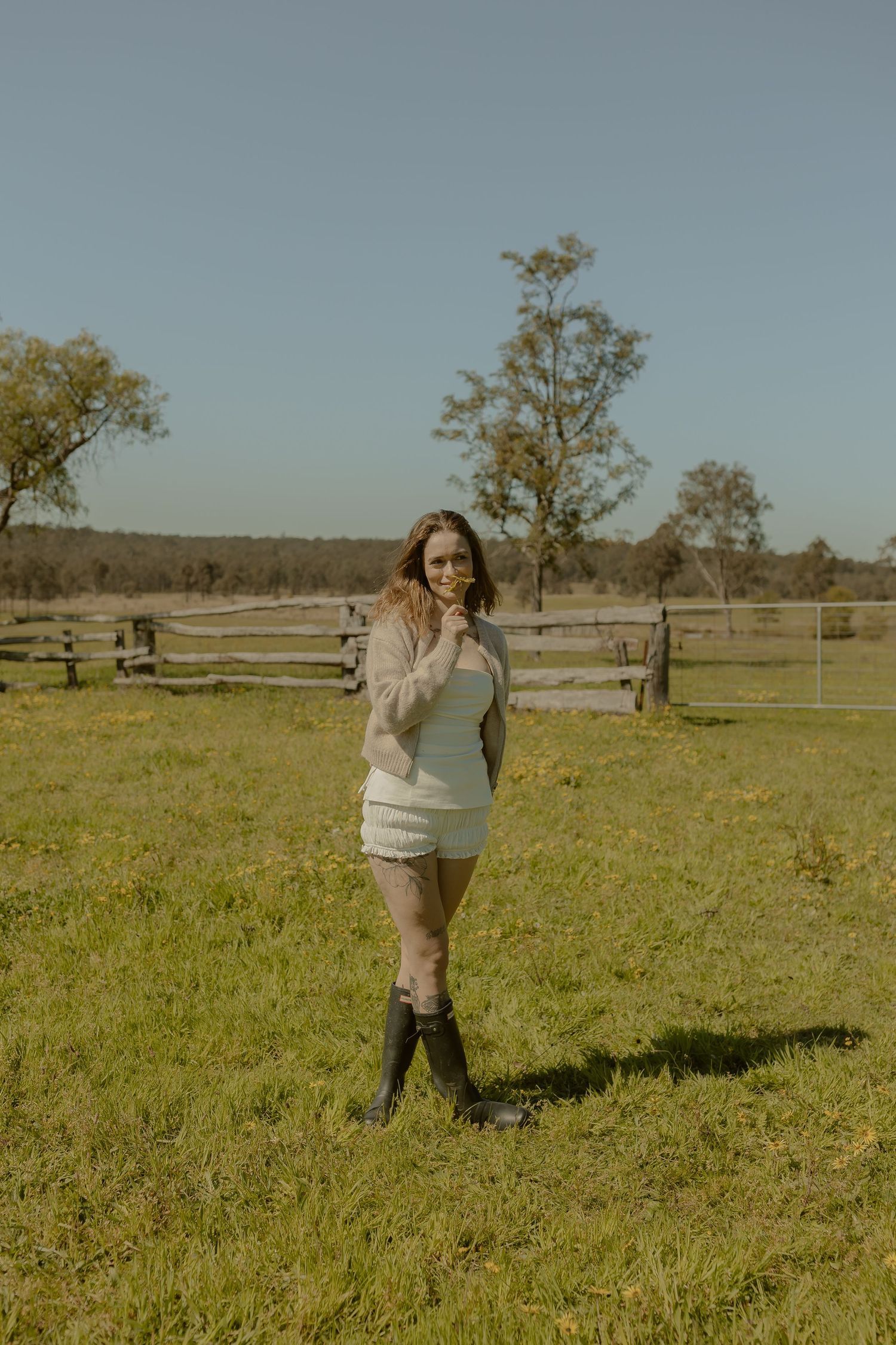 A person in a light sweater walks through a grassy field with wooden fences and trees in the distance.