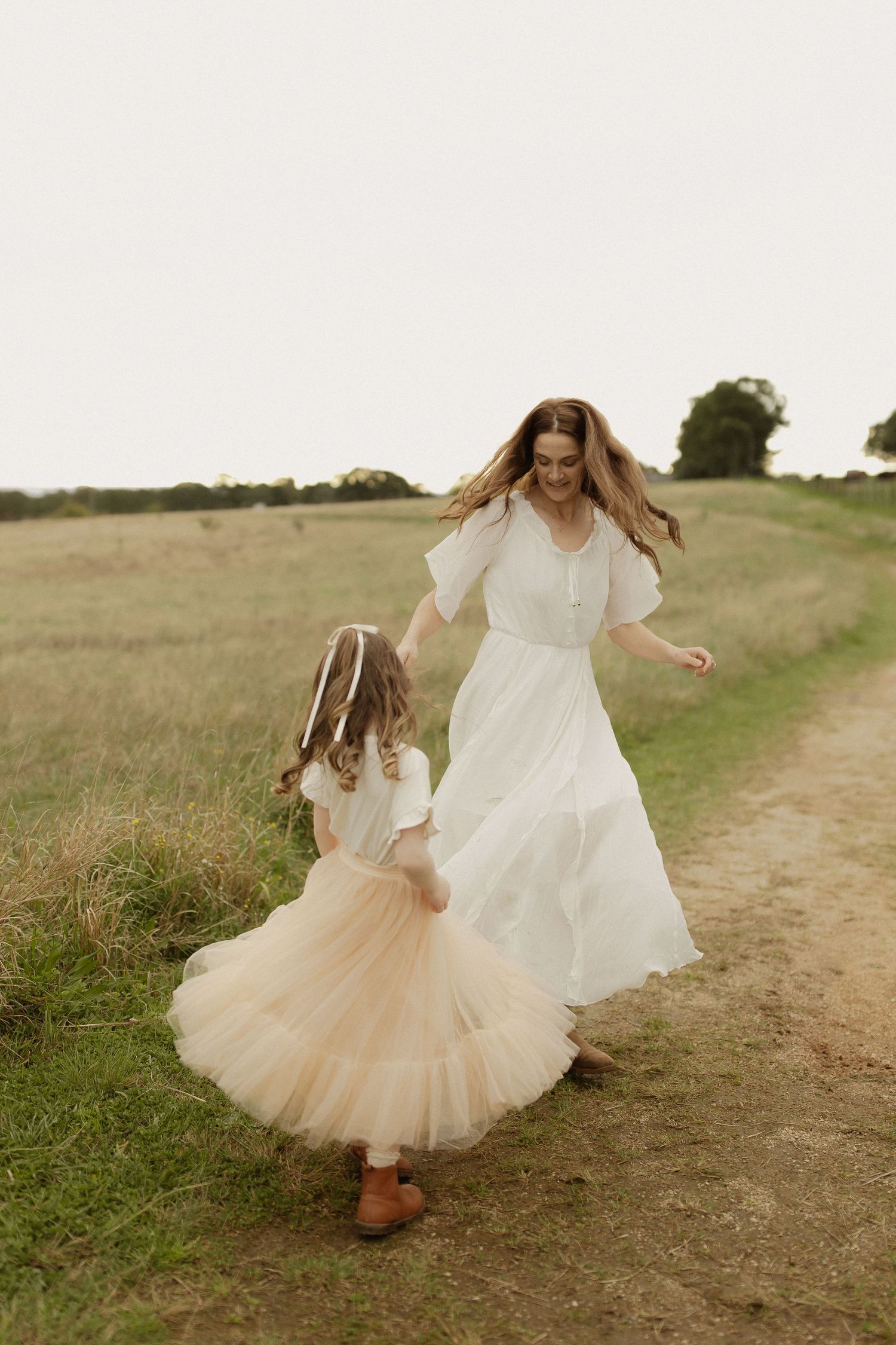 Two figures in contrasting white and peach dresses twirl together on a rustic countryside path.