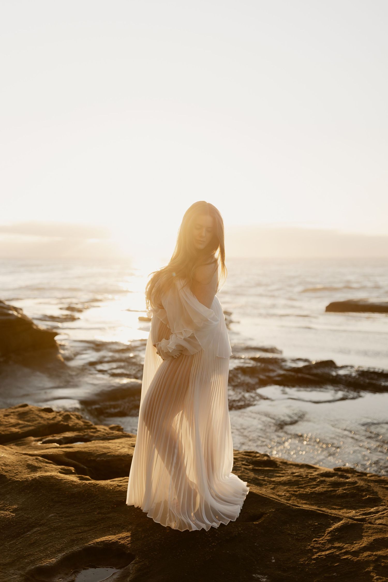 Silhouetted maternity figure in flowing white dress stands on rocky coastline at sunset with ocean waves crashing below.