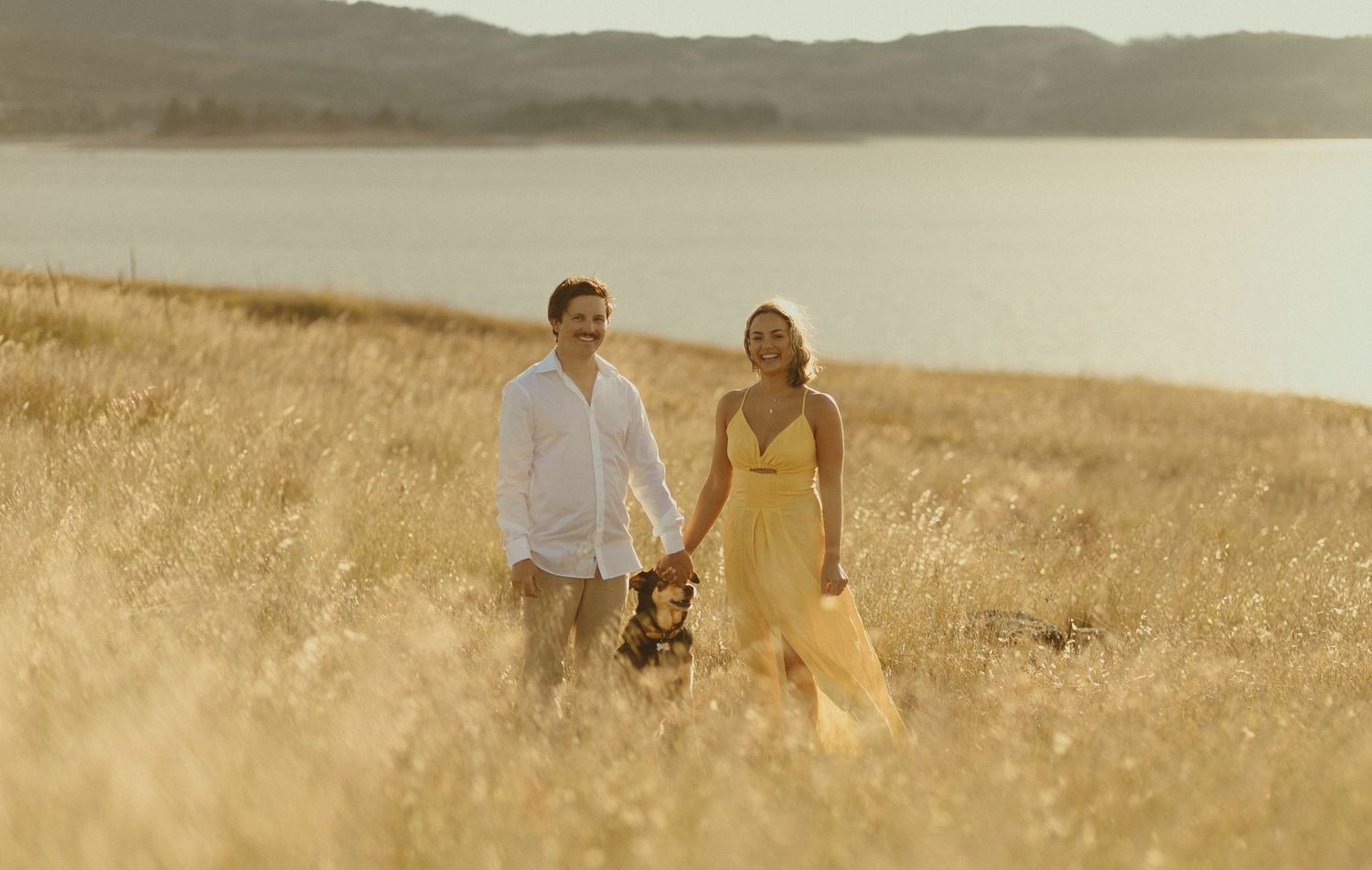Two people walk hand in hand through a golden sunlit field beside a serene lake at sunset.