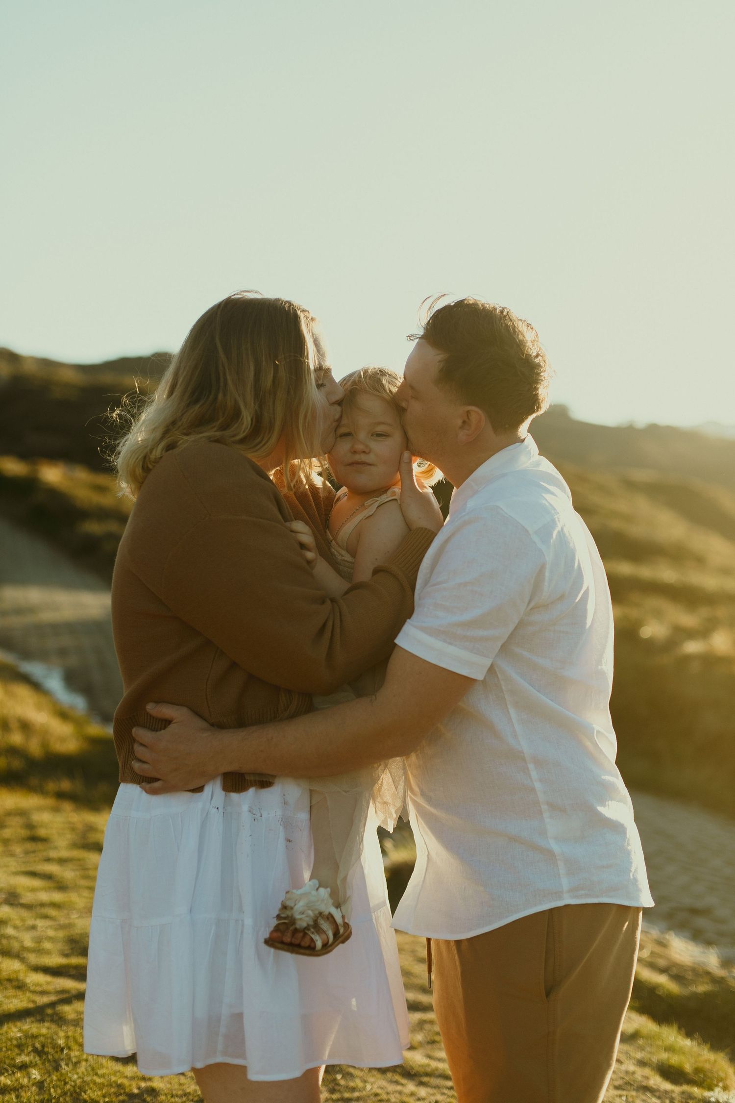 A family shares a tender moment together during golden hour on a grassy hillside at sunset.