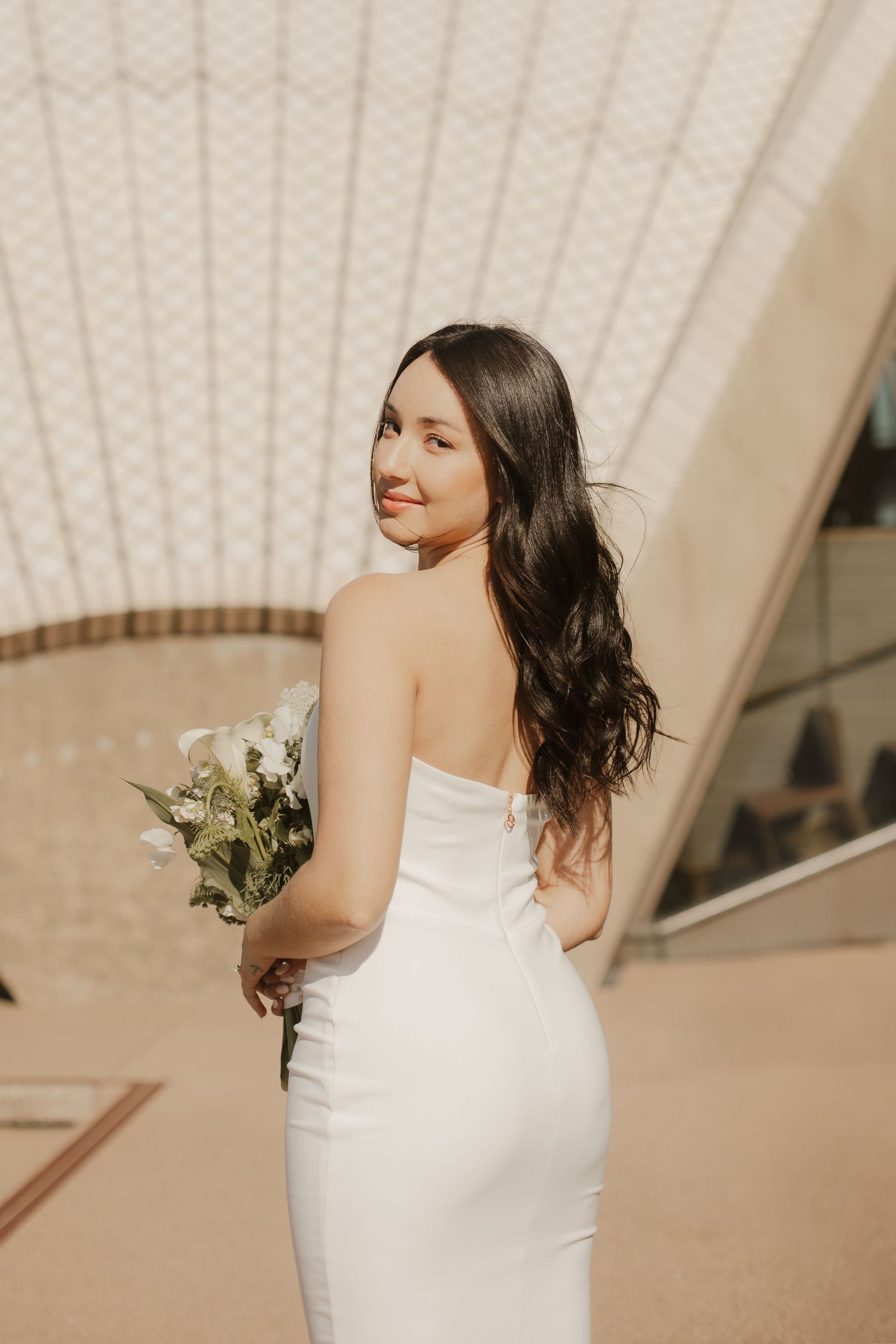 Bride in sleek white wedding dress holds bouquet while looking over shoulder in warm interior setting.
