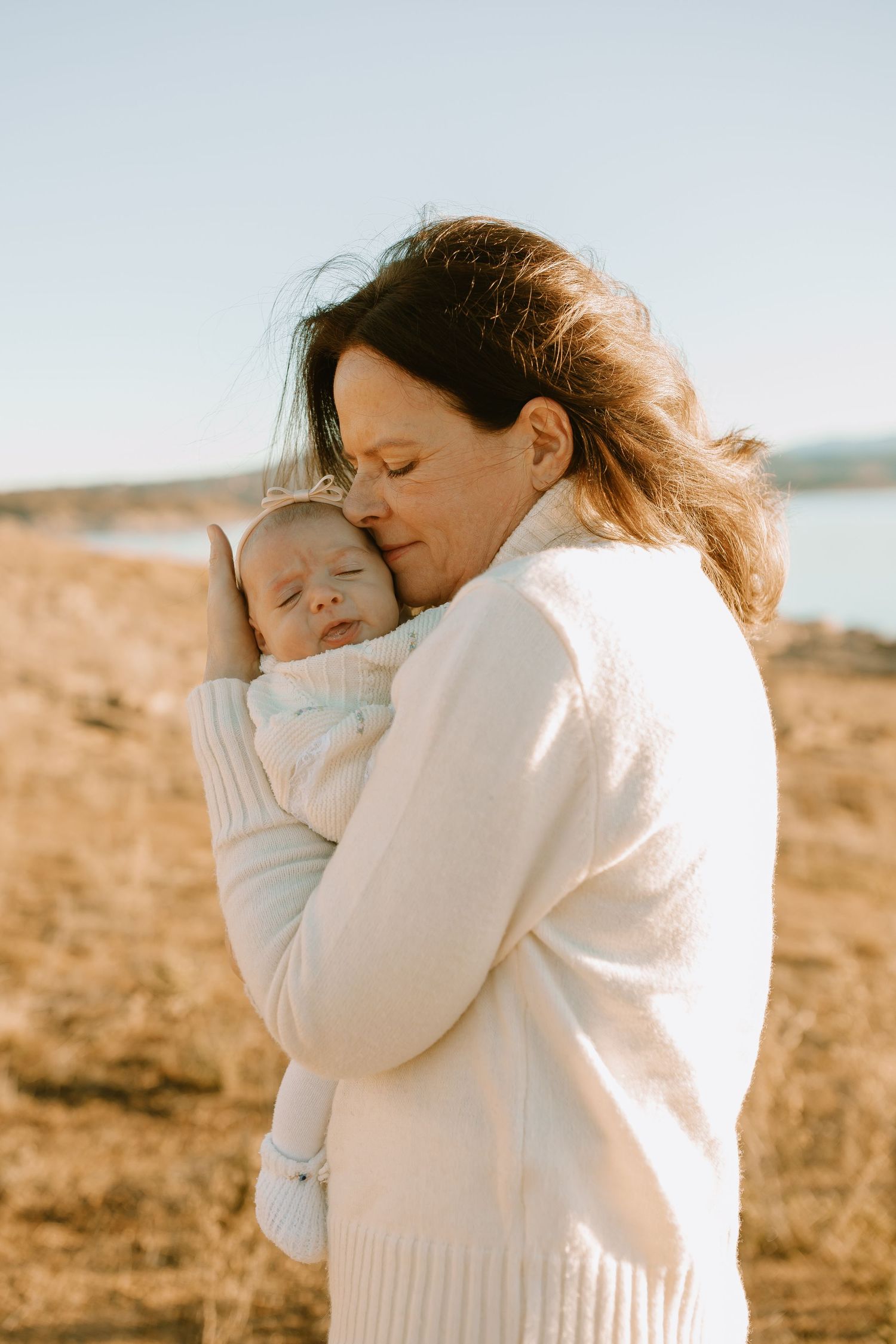 Intimate outdoor portrait of someone in white sweater embracing infant against coastal landscape backdrop.