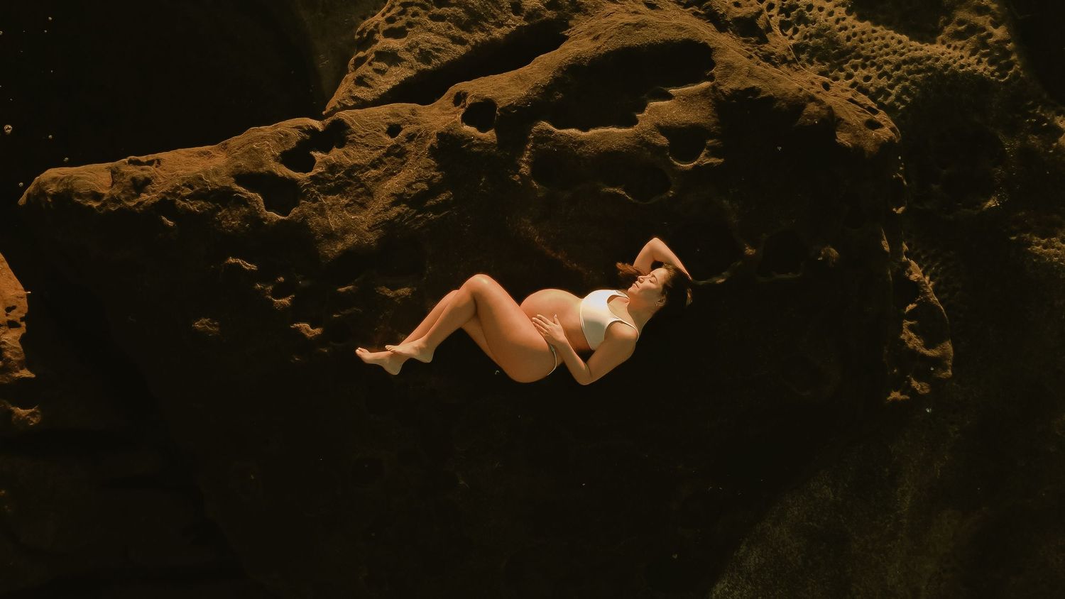 Person in white swimwear reclines gracefully against dramatic rock formations at golden hour by the ocean.