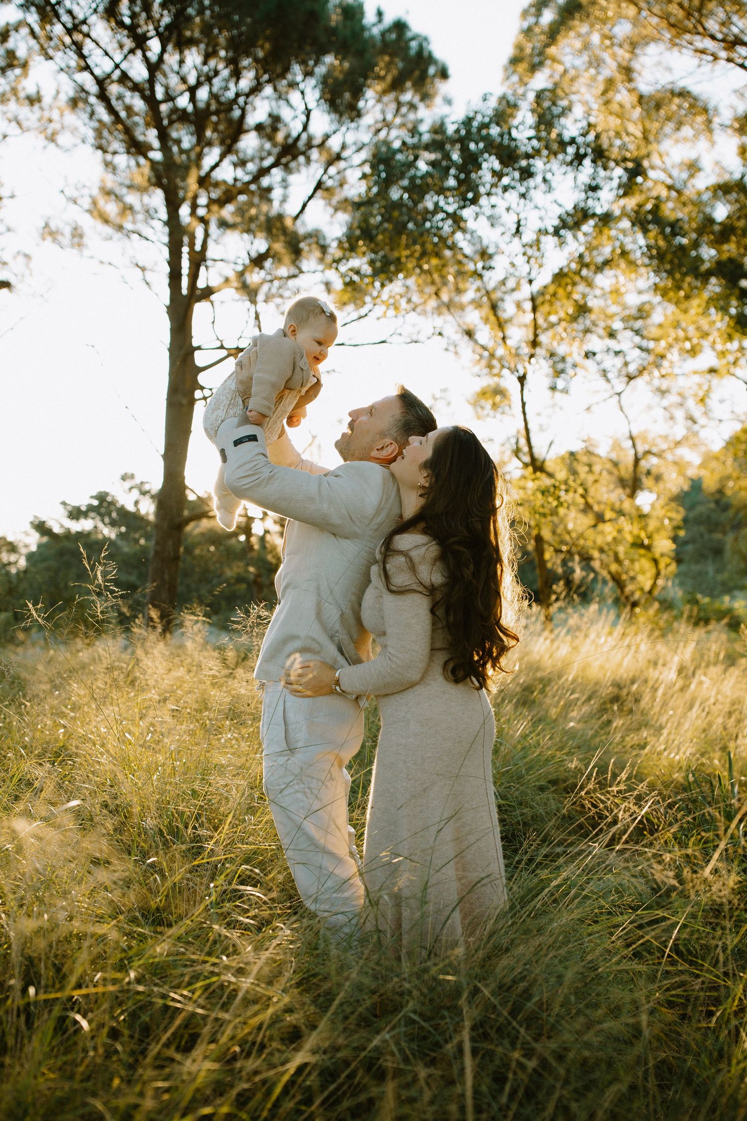Family enjoying sunset moment as parents lift baby in grassy field.