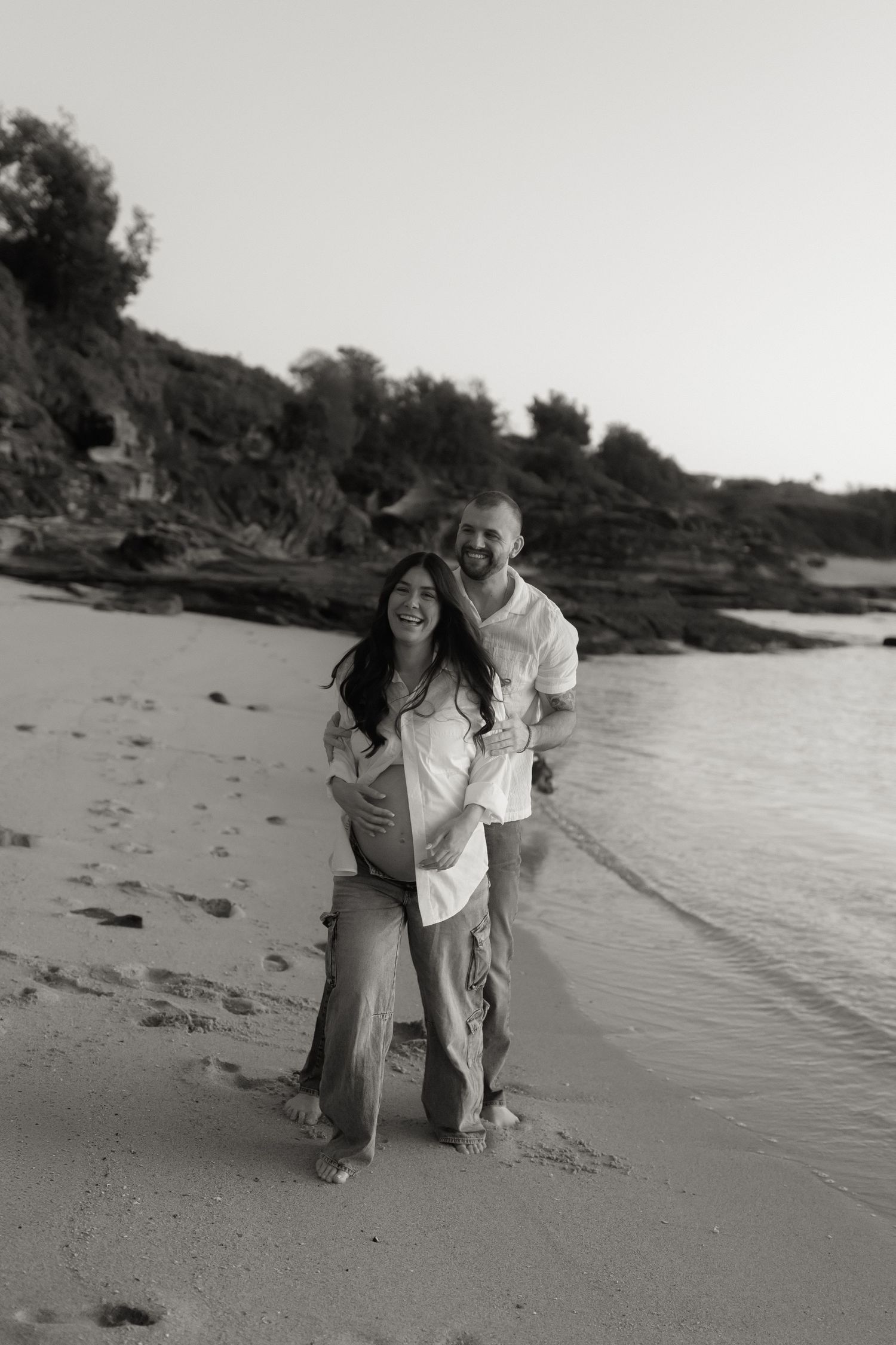 Black and white photo of a happy couple on a beach, man embracing pregnant woman from behind, rocky cliffs in background.