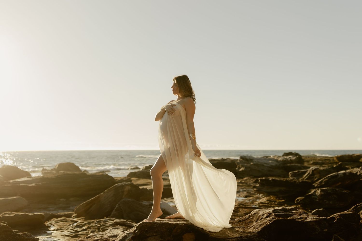 Pregnant woman in flowing white dress standing on coastal rocks at sunset with ocean waves in background.