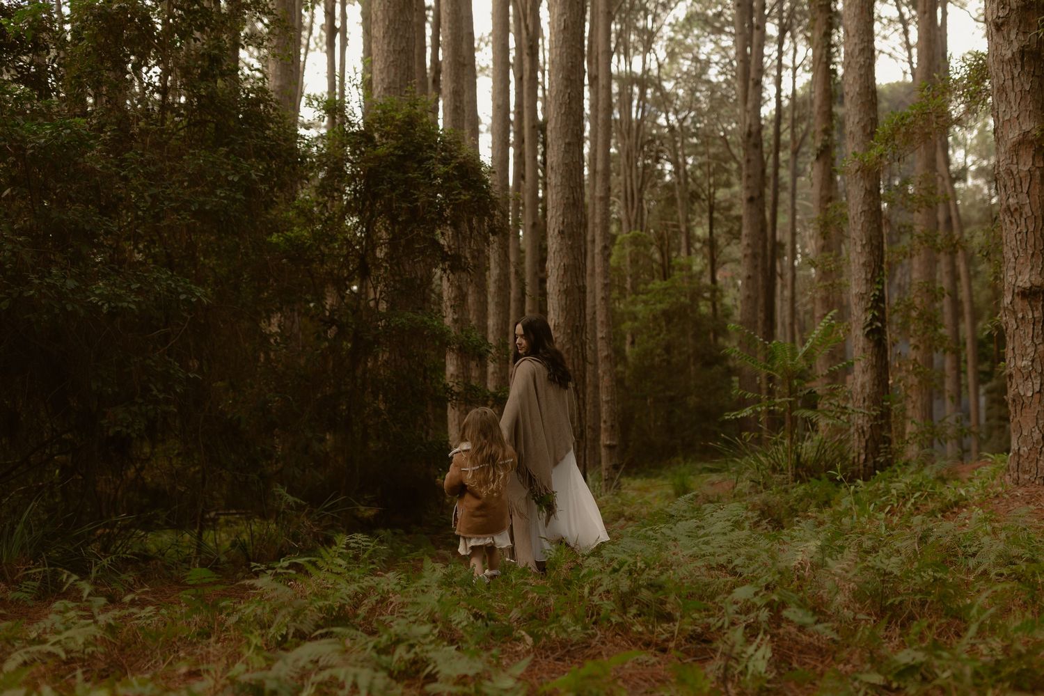 Woman in long grey dress walking with young child through lush green forest path.