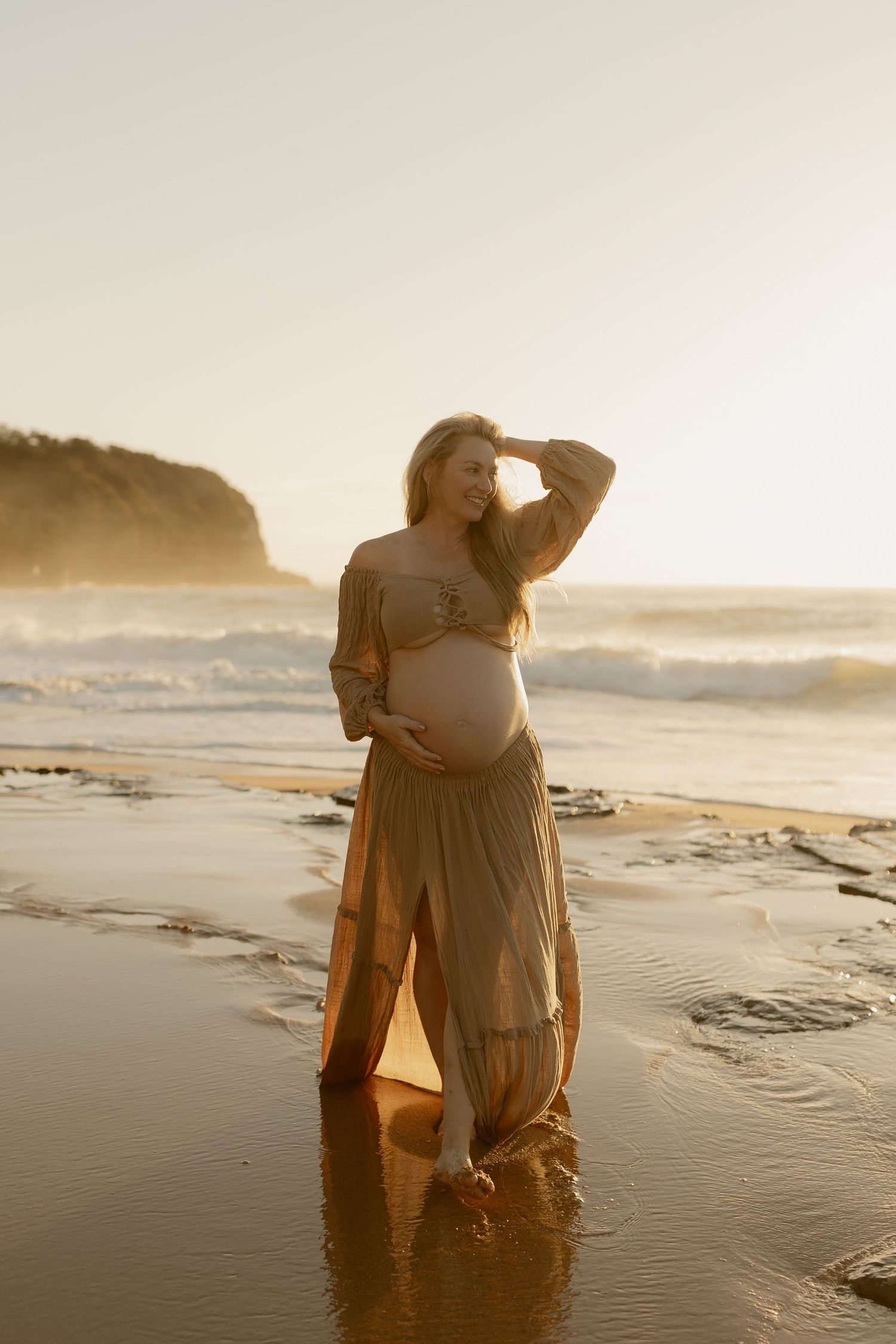 Glowing pregnant woman in a flowing olive dress standing in beach waves during a stunning sunset.