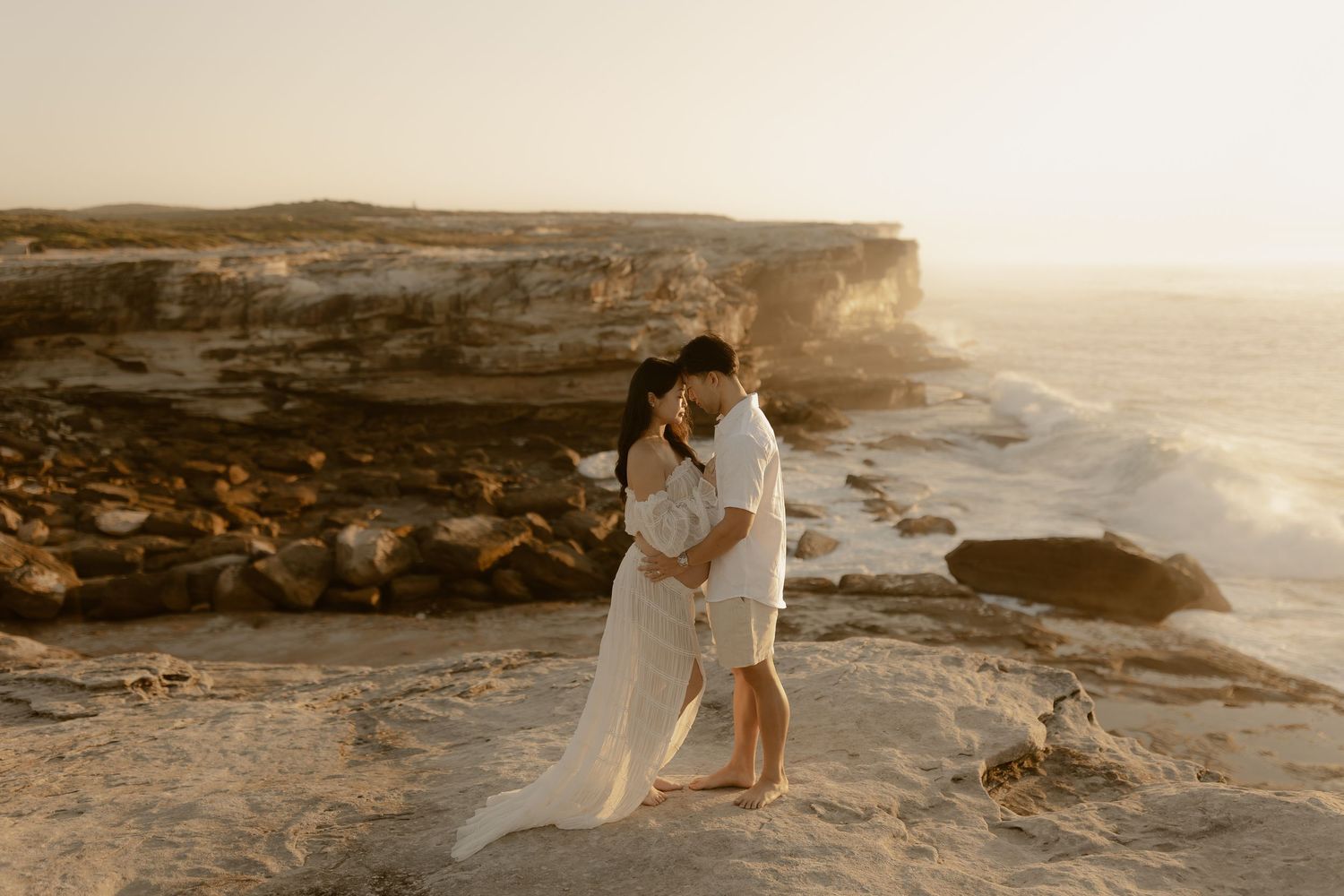 A couple in wedding attire embraces on a rocky coastal cliff at sunset creating a romantic silhouette.
