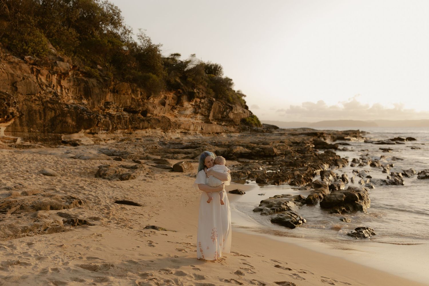 A couple embraces on a secluded rocky beach at sunset with cliffs and ocean waves in the background.