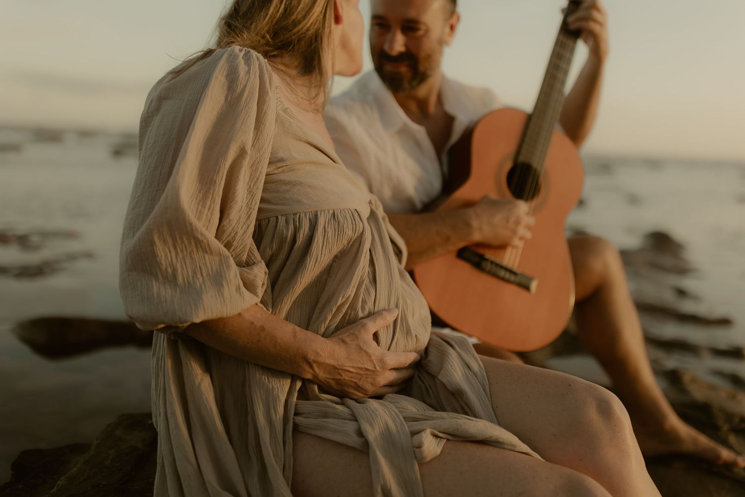 Couple in light flowy clothing sits intimately on beach at sunset while playing acoustic guitar.