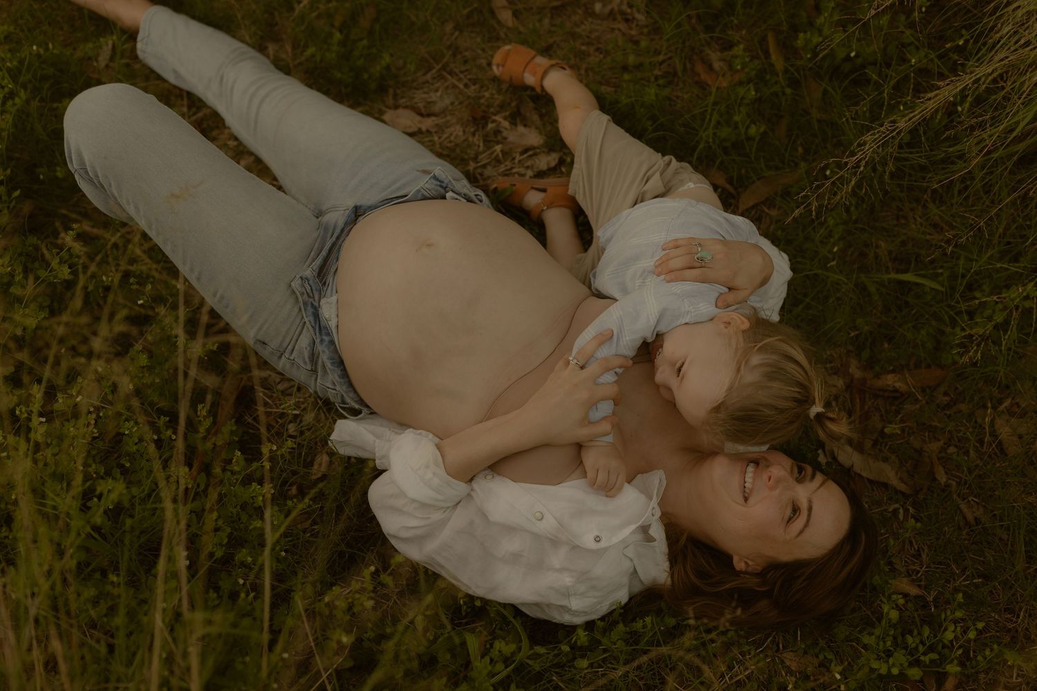 Pregnant woman lying in grass with young child resting on her baby bump during outdoor maternity photo session.