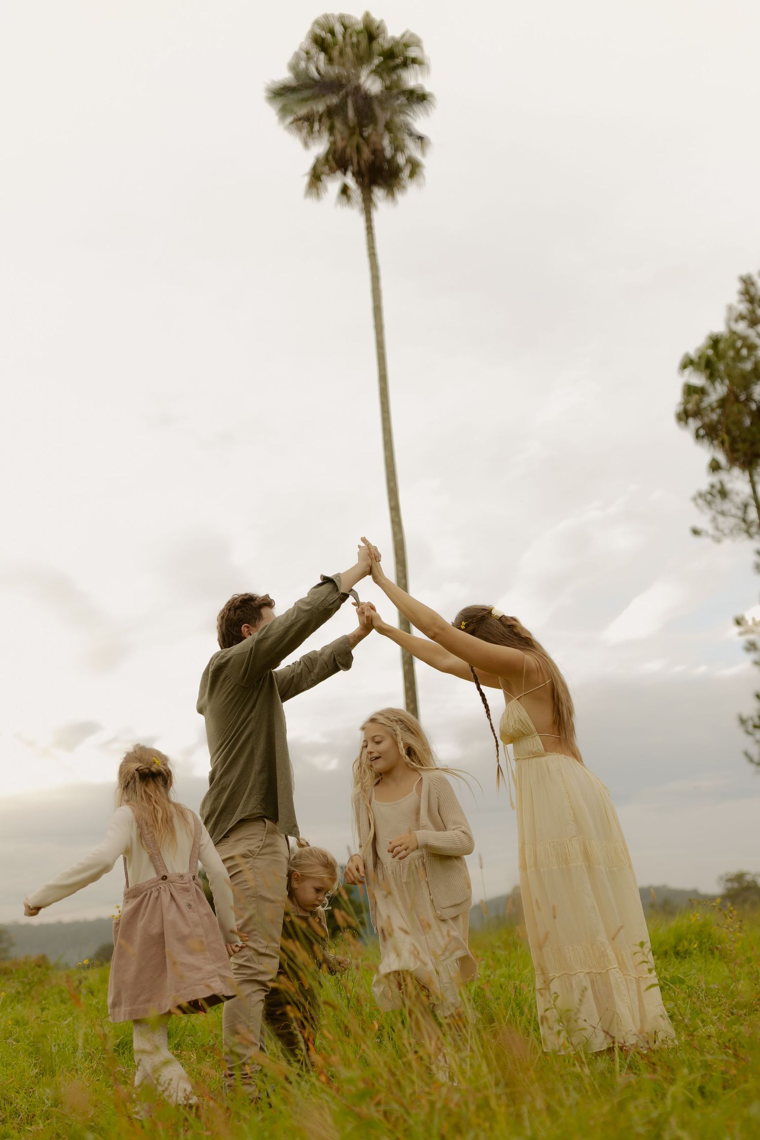 Family of five joyfully raising hands together in a grassy field with a tall palm tree under an overcast sky.