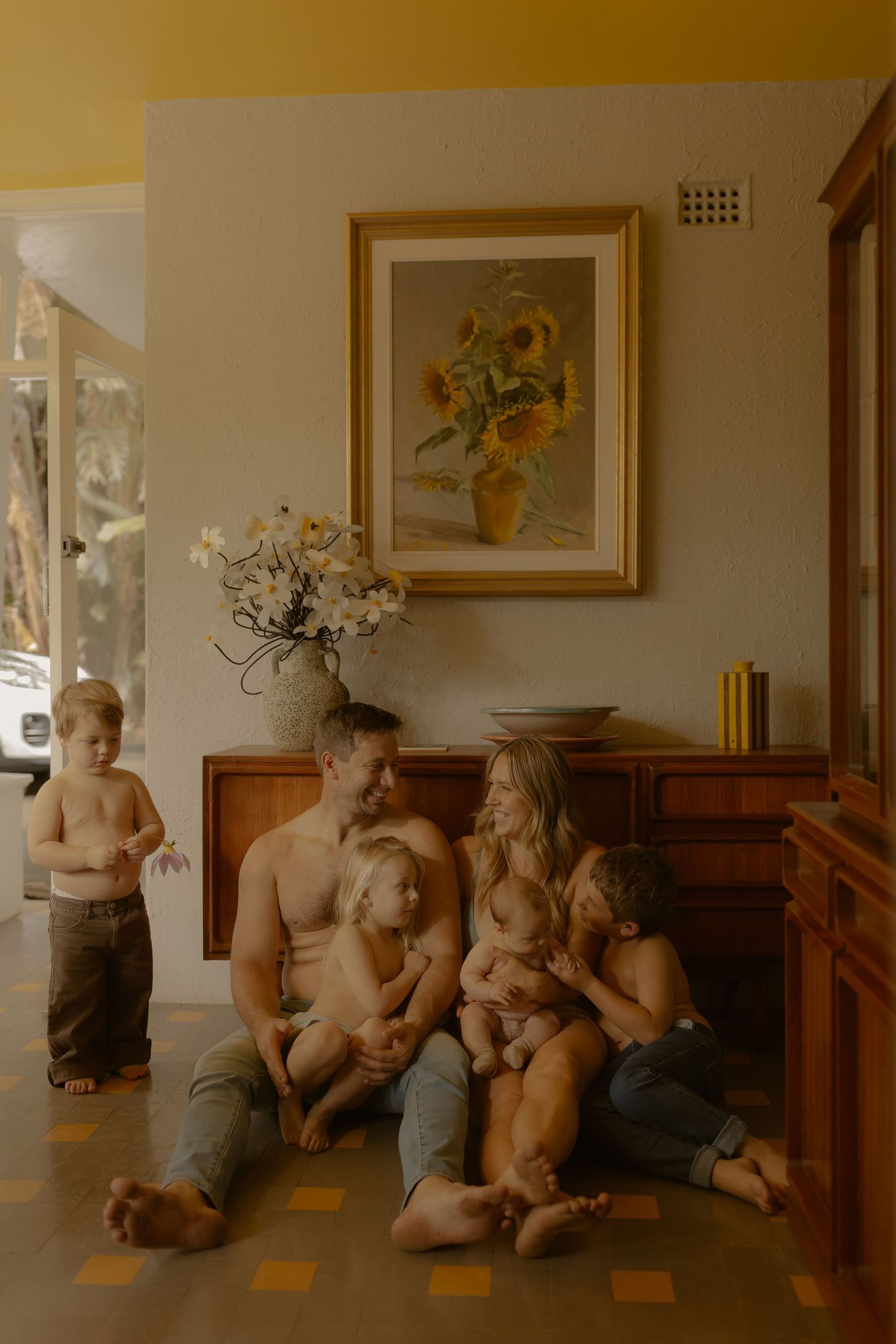 A happy family of five sits together on the floor beneath a sunflower painting in a warm, golden-toned home.