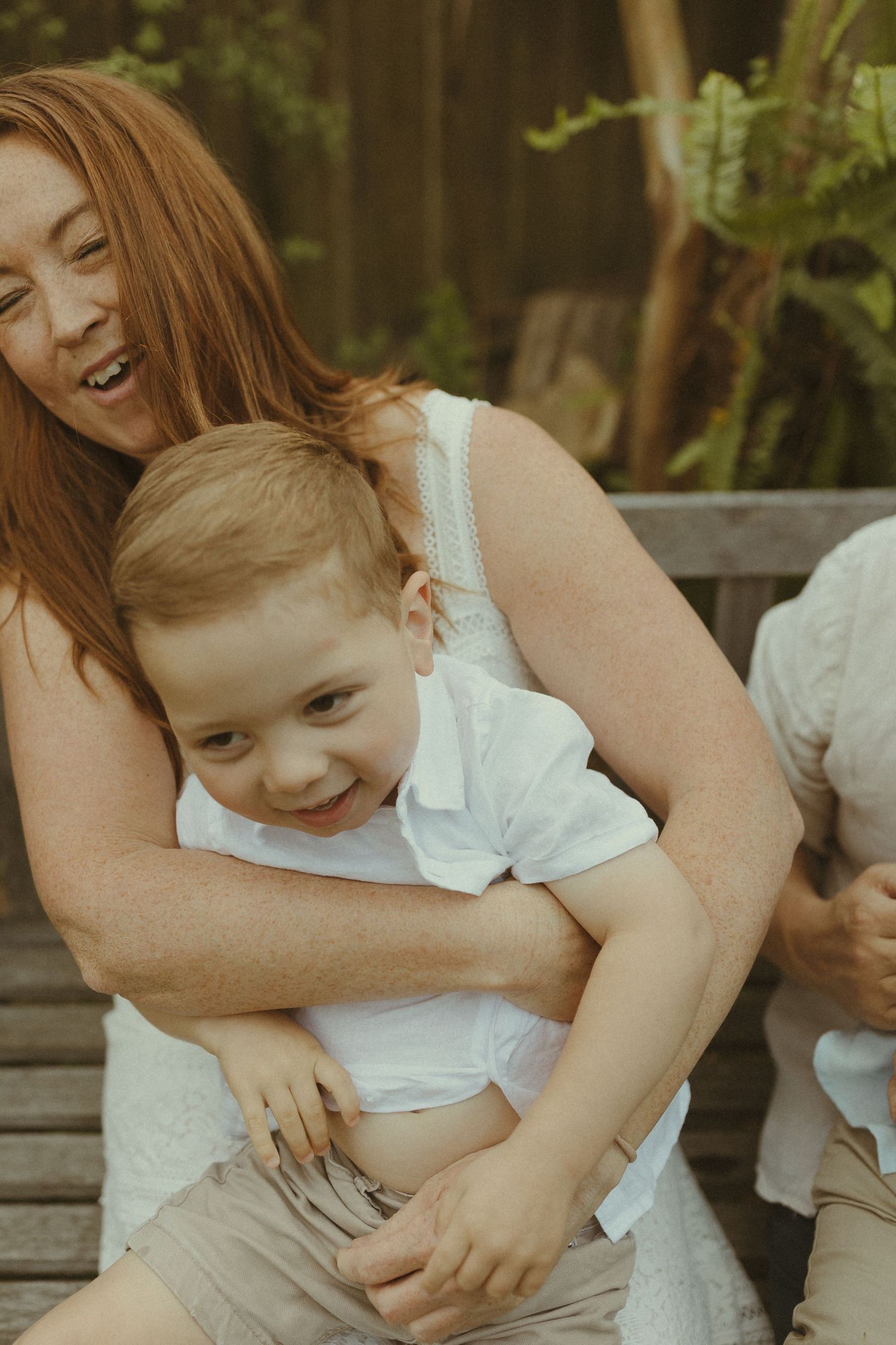 A playful moment of laughter and cuddles between parent and child on a wooden bench outdoors.