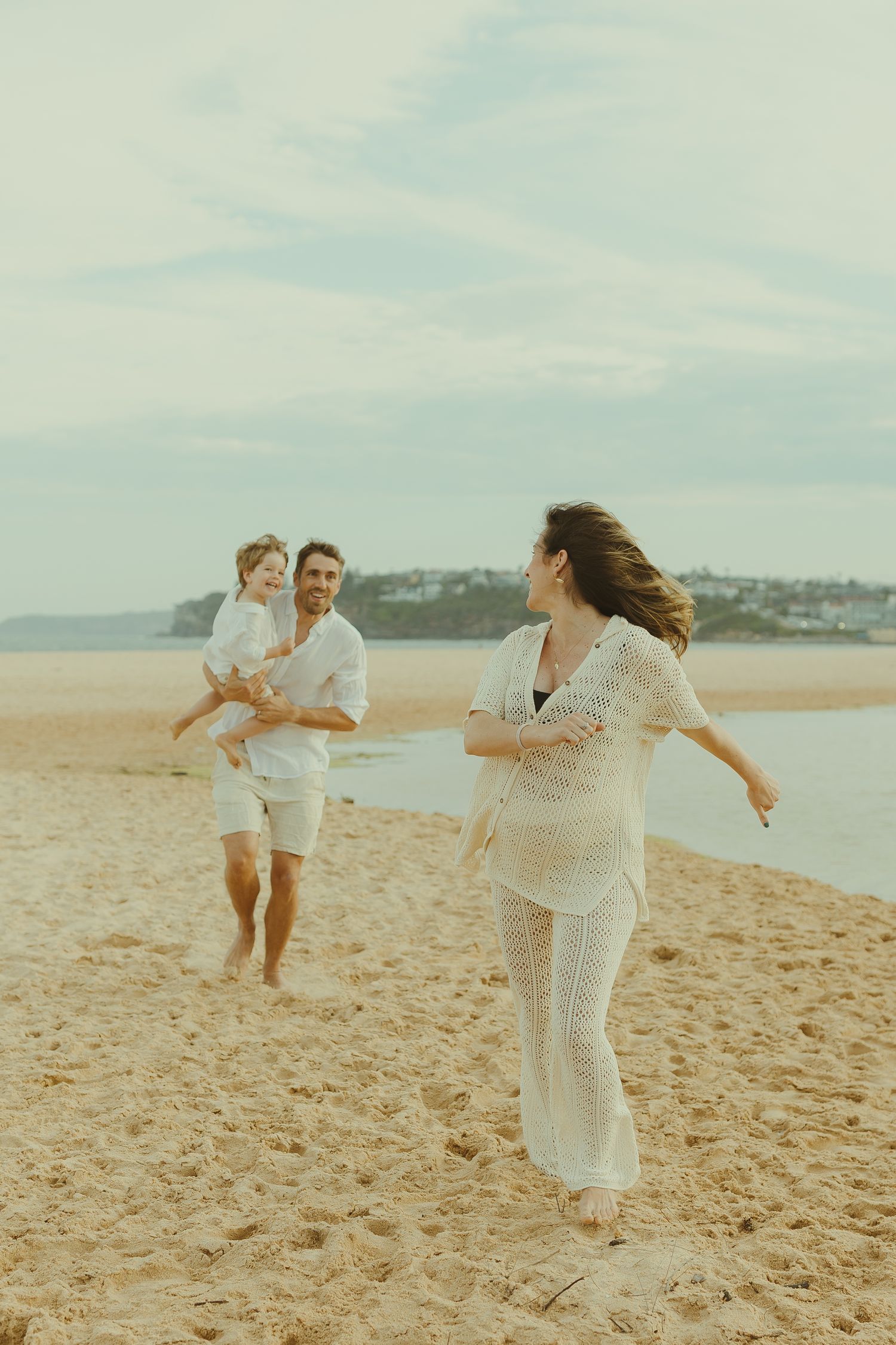 Family playing and running together on a sunny beach with coastal views in the background.
