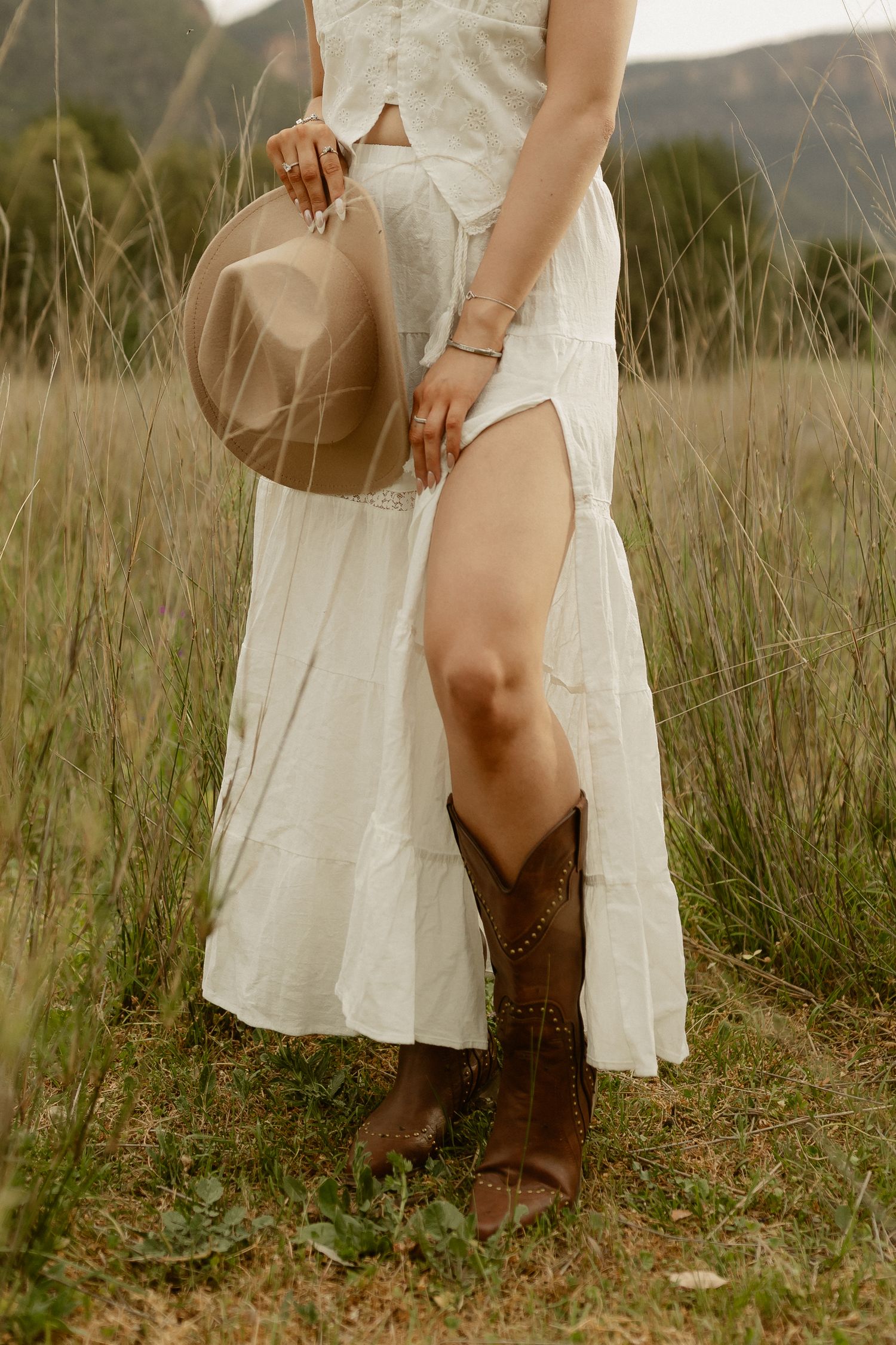 Woman in white dress holding tan hat and wearing brown cowboy boots in rustic outdoor setting.
