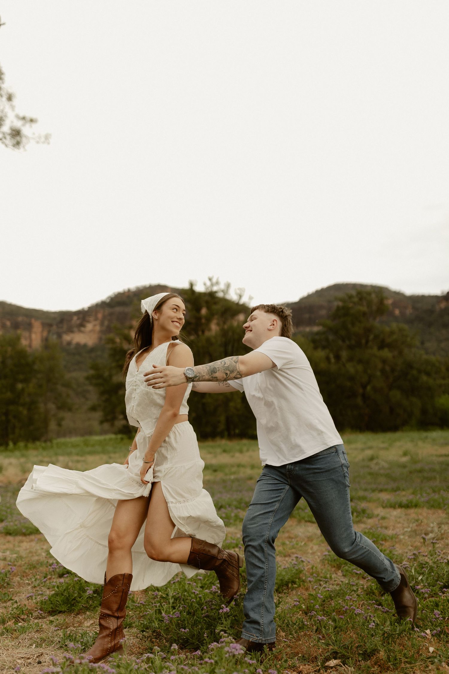 Playful couple dancing and twirling in a field with mountains visible in the distance.