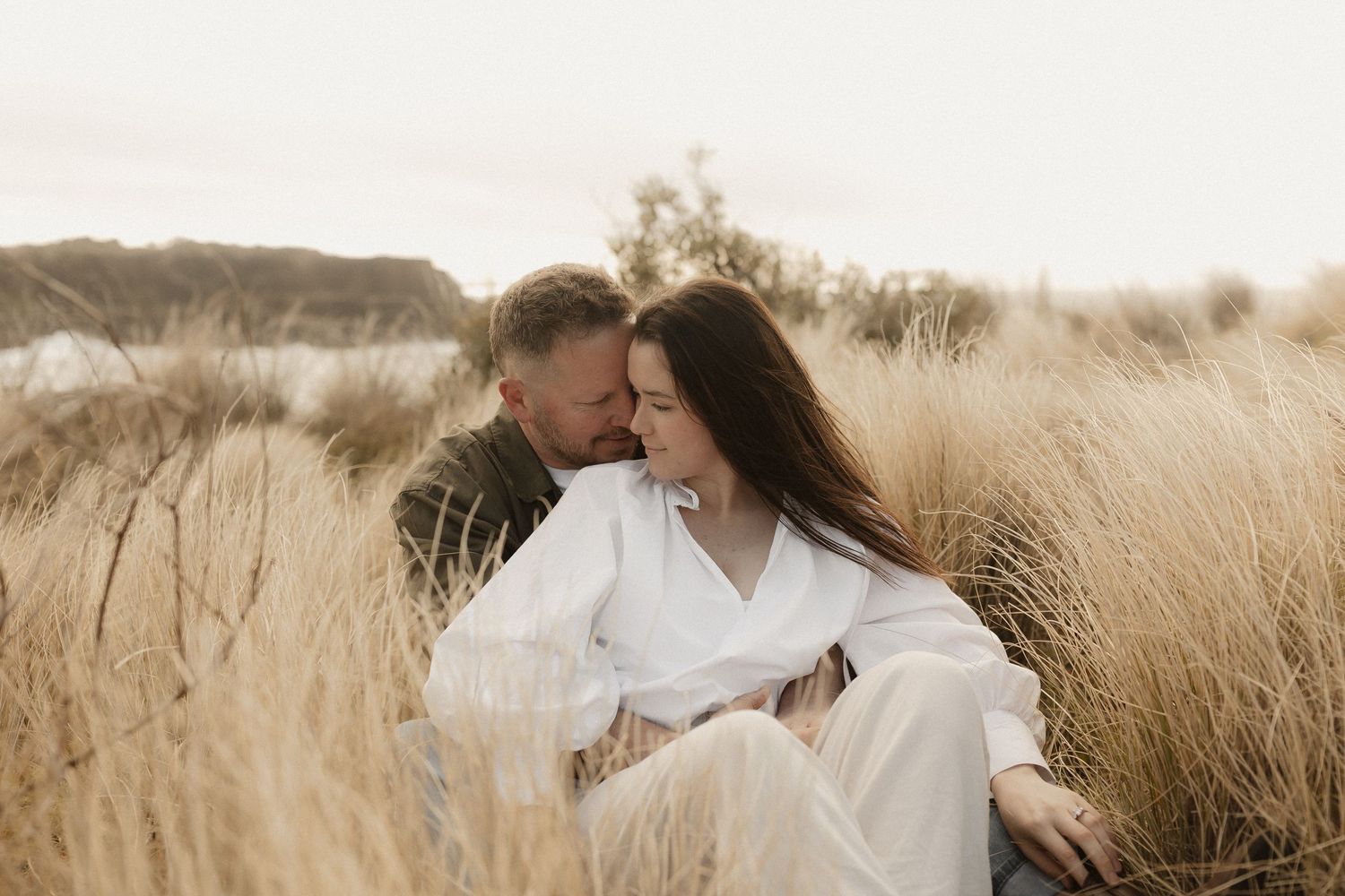 Romantic couple embraces in tall golden grass field during sunset creating dreamy ethereal atmosphere.