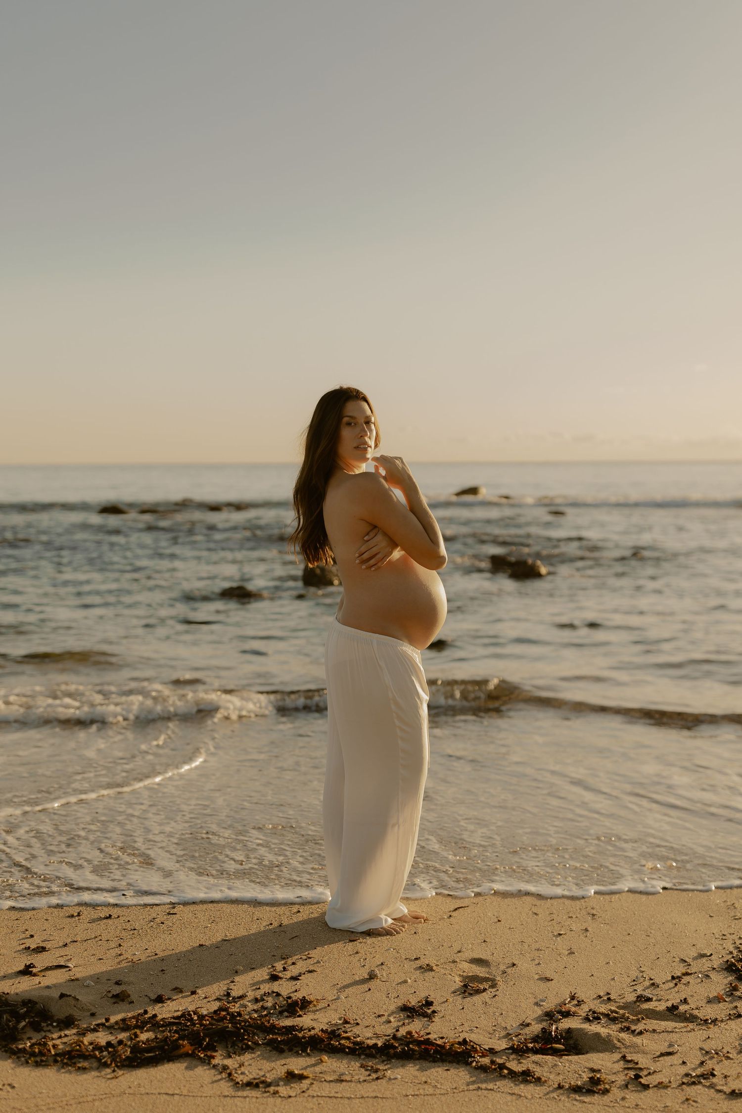 A person in white attire poses elegantly on a sandy beach with ocean waves in the background.