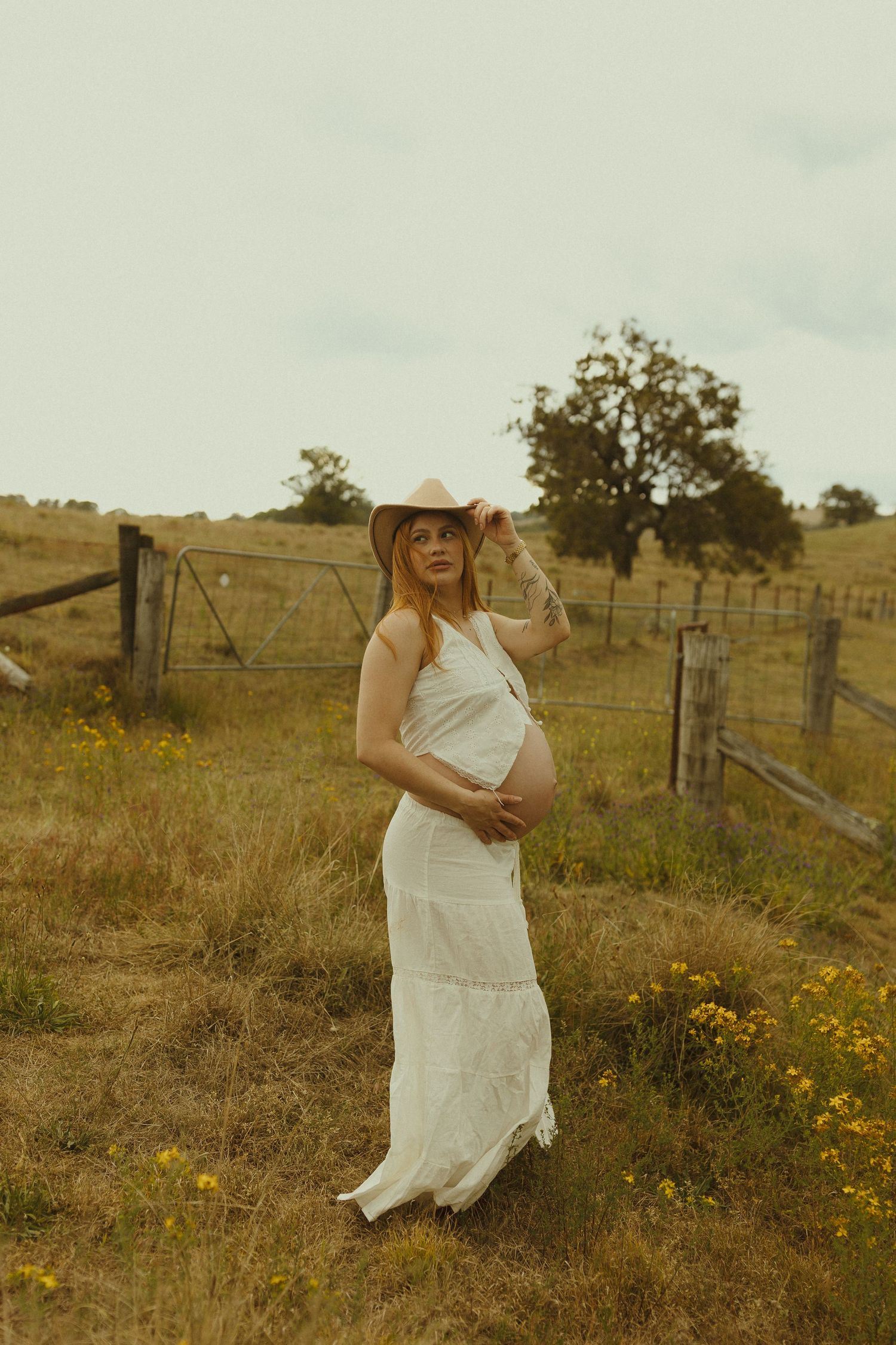 A serene maternity portrait in a white dress and hat against a rustic farm setting.