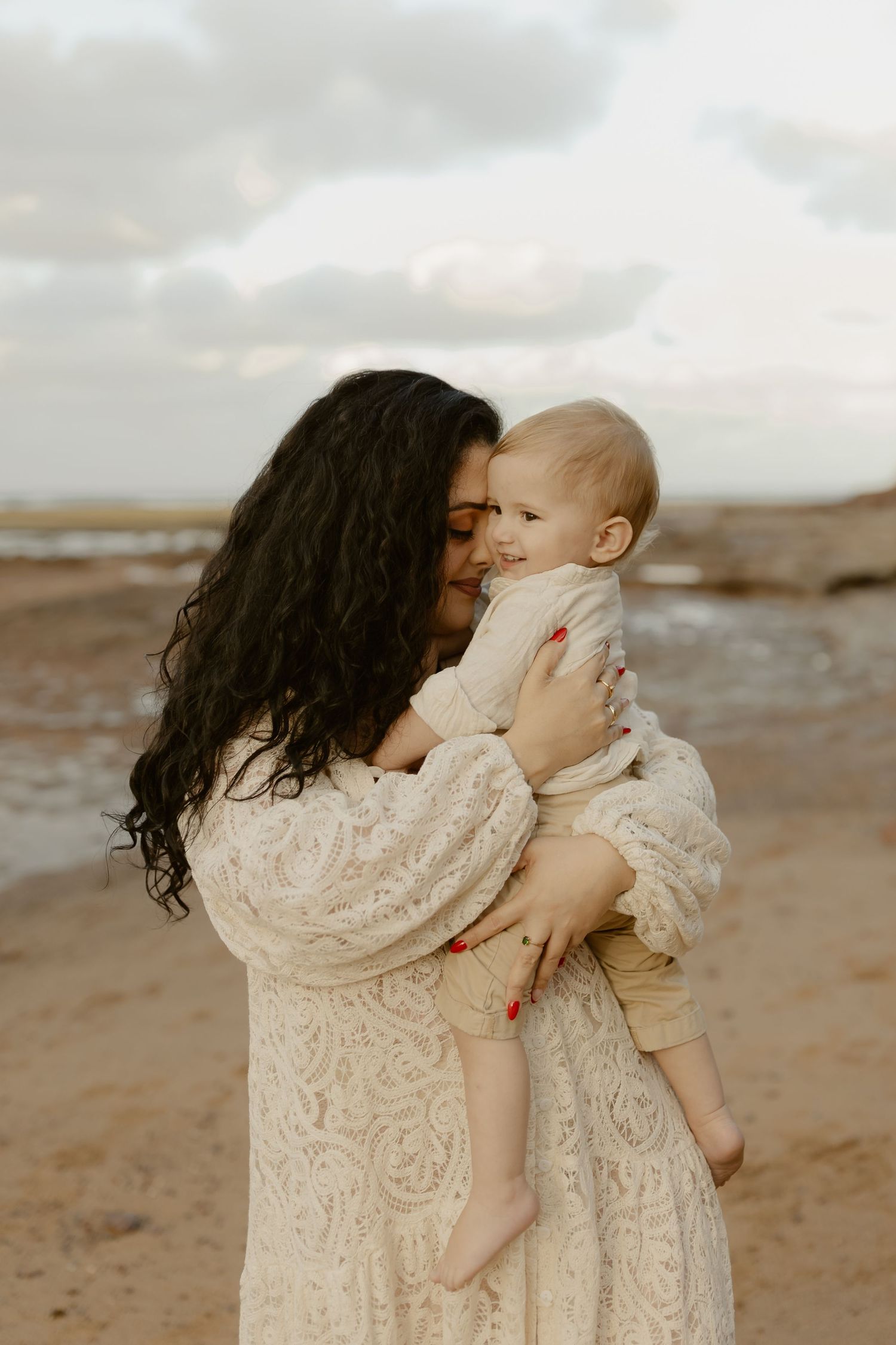 A tender moment captured on a beach between parent and child wearing matching cream lace dresses at sunset.